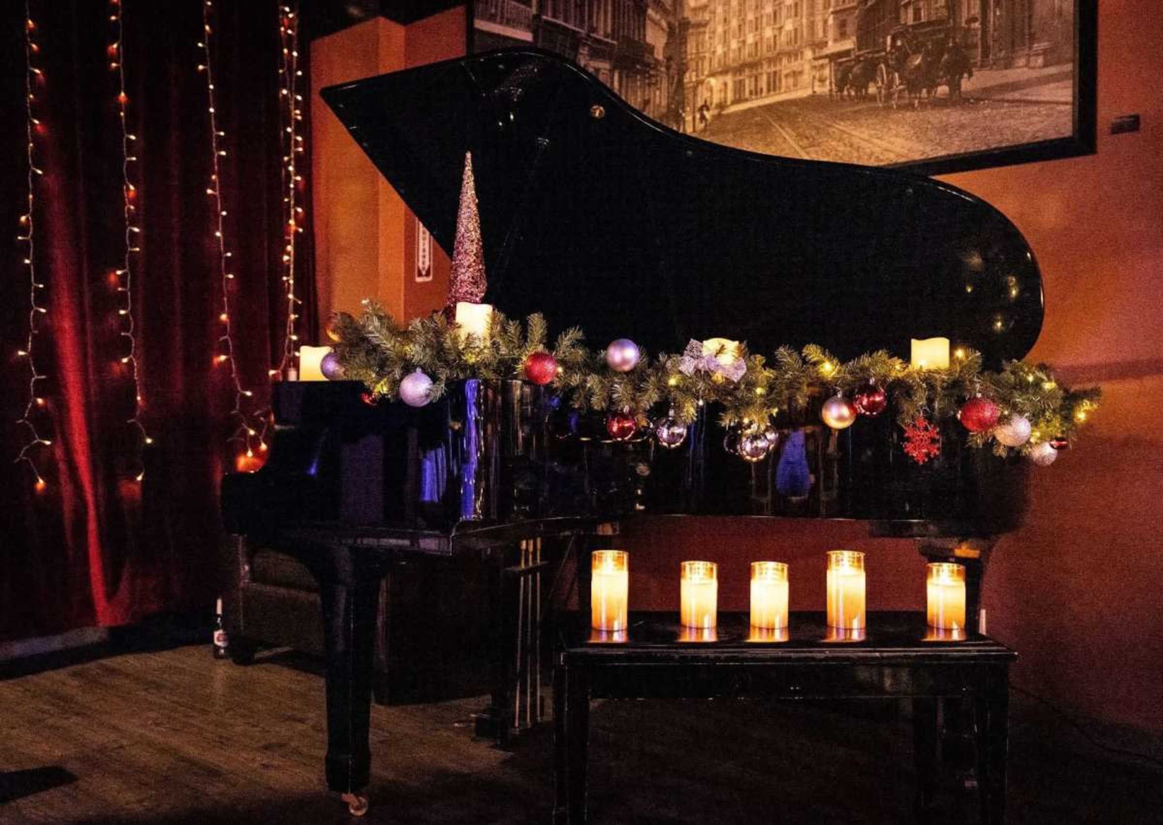 A grand piano decorated with garlands, ornaments, and candles stands against a backdrop of red curtains and vintage photographs.