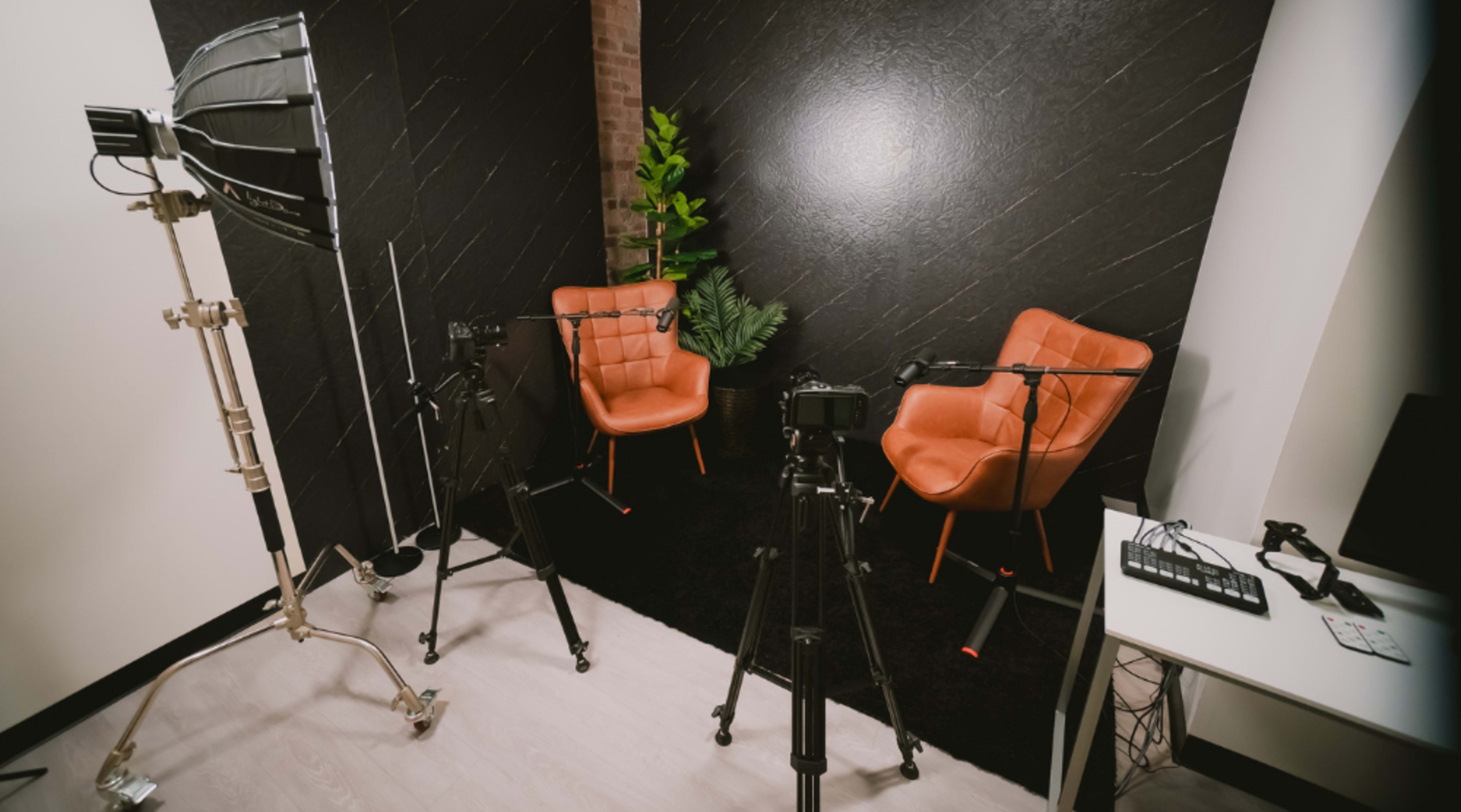 The image shows a small studio setup with two orange chairs facing each other, surrounded by cameras and lighting equipment, against a dark textured wall.