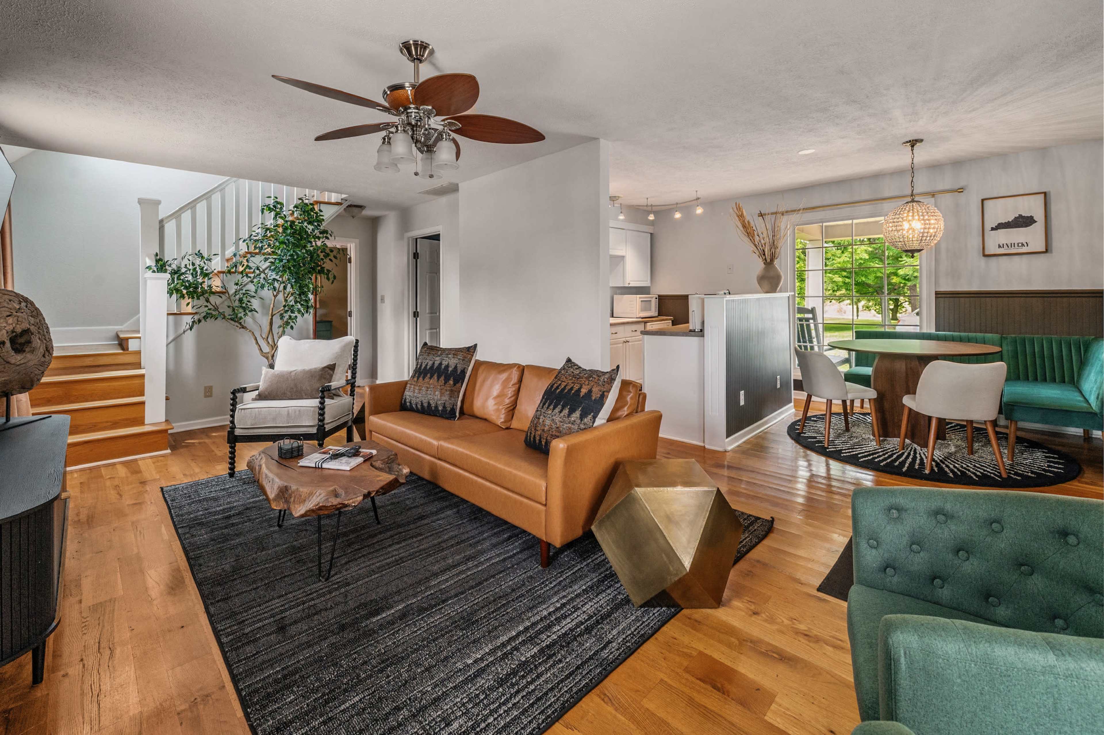 The image shows a spacious living area featuring a light brown leather sofa, a dining table, and a staircase leading to another level, with large windows providing natural light.