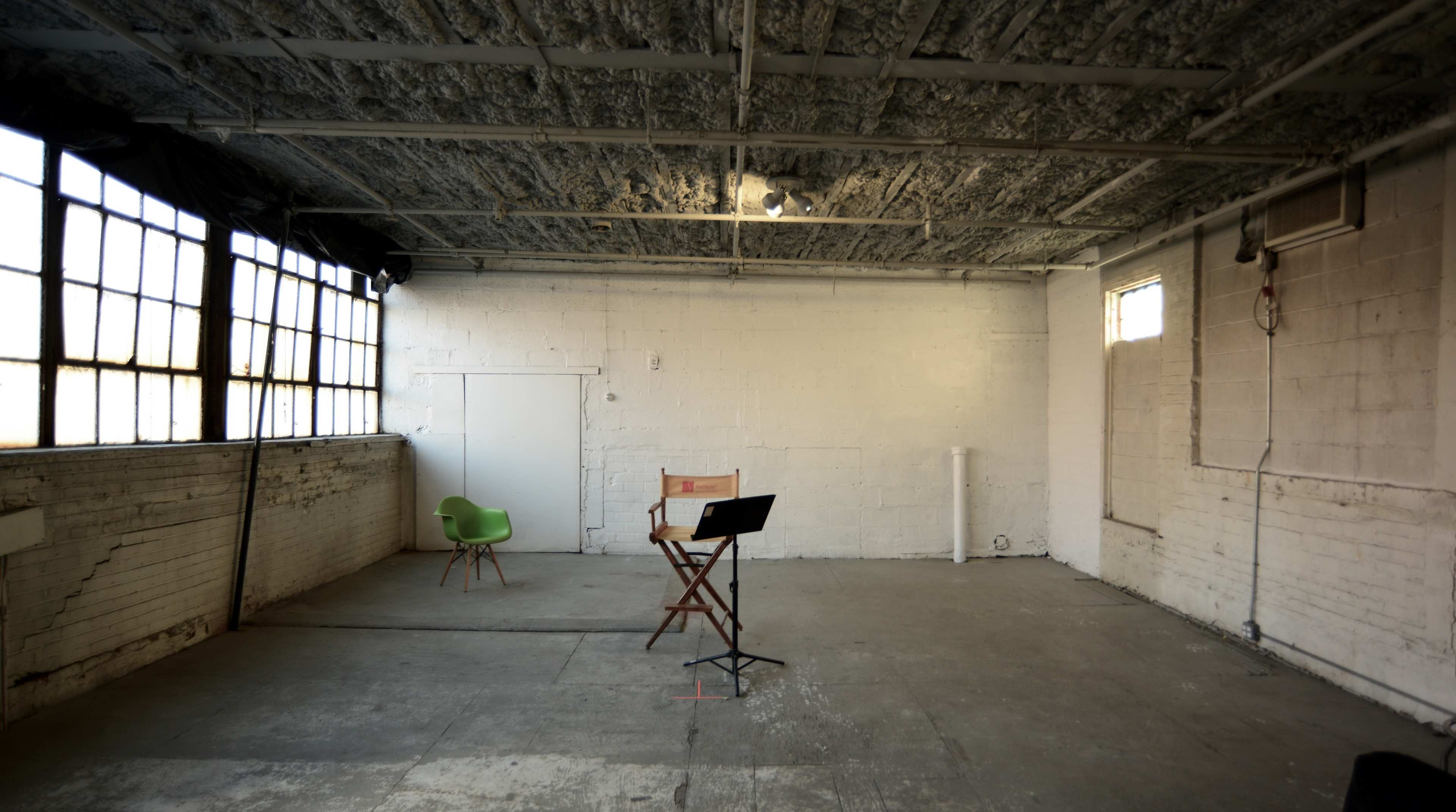 An empty industrial room with a wooden chair, a green chair, and a music stand on a concrete floor.
