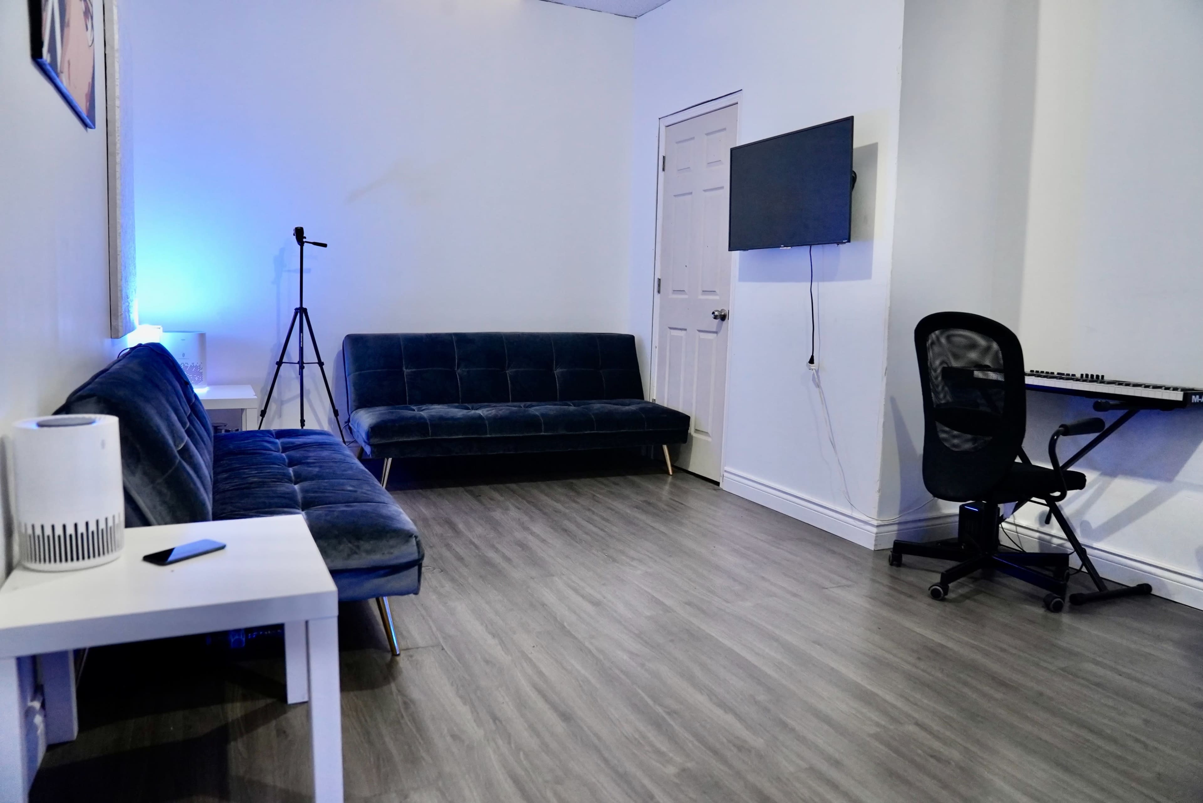 A minimalist room features two blue sofas, a small white table, a microphone stand, a wall-mounted TV, and a black office chair next to a keyboard.