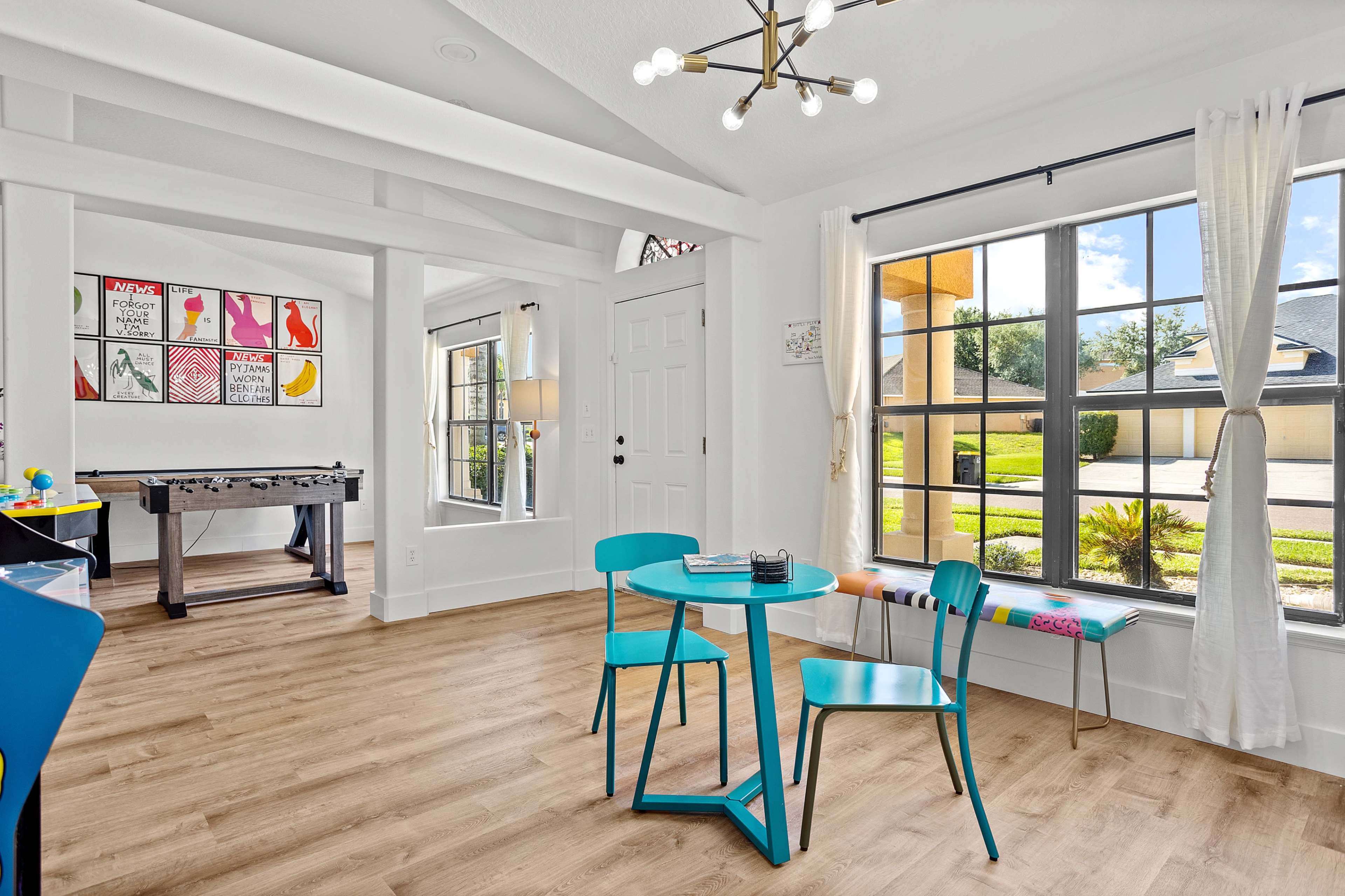 A brightly lit room features a small round blue table and two matching chairs, with a foosball table in the background and large windows that open to an outdoor view.