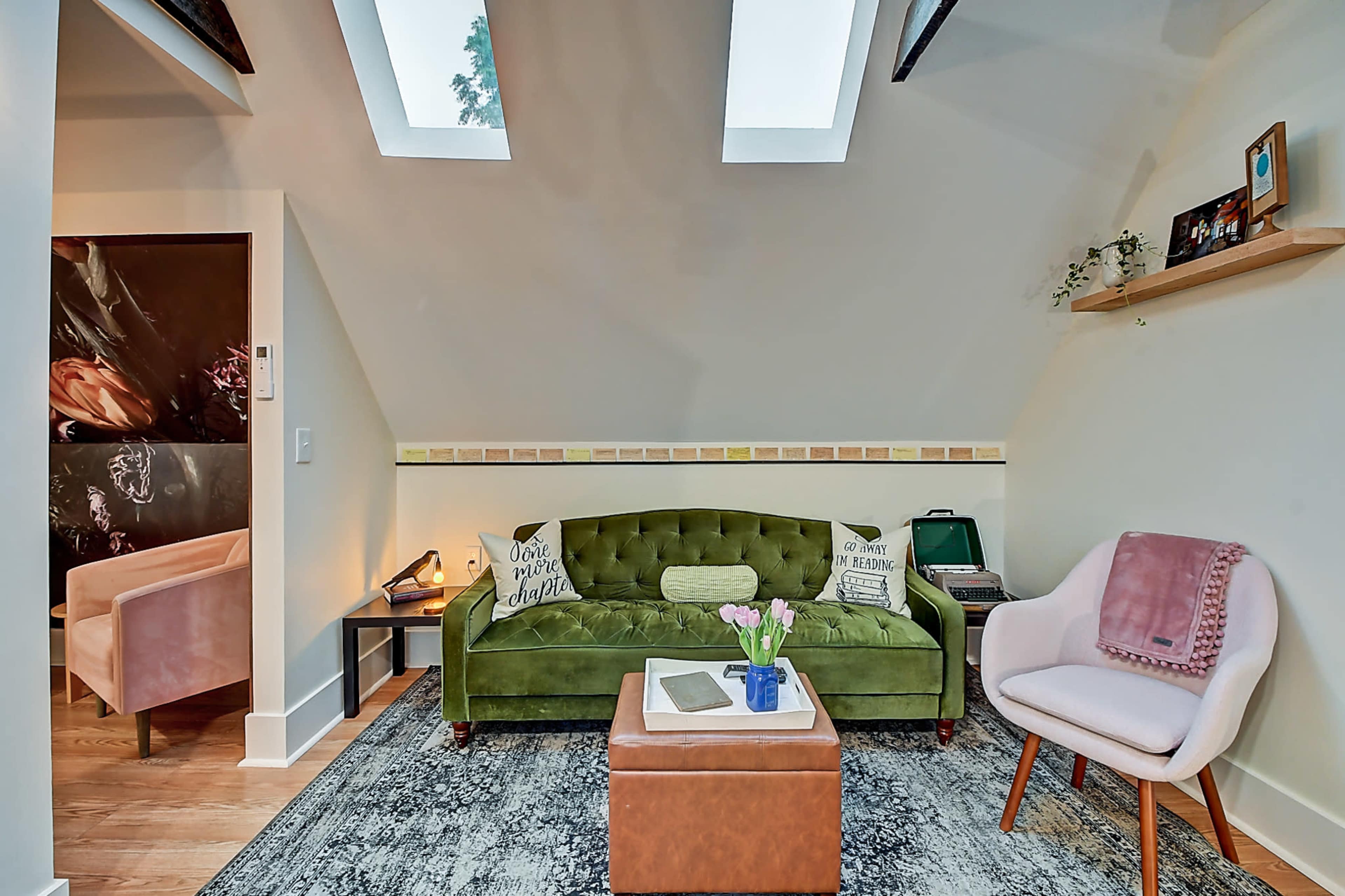 A cozy living room features a green velvet sofa, a pink armchair, and a brown coffee table, complemented by skylights and a colorful accent wall.
