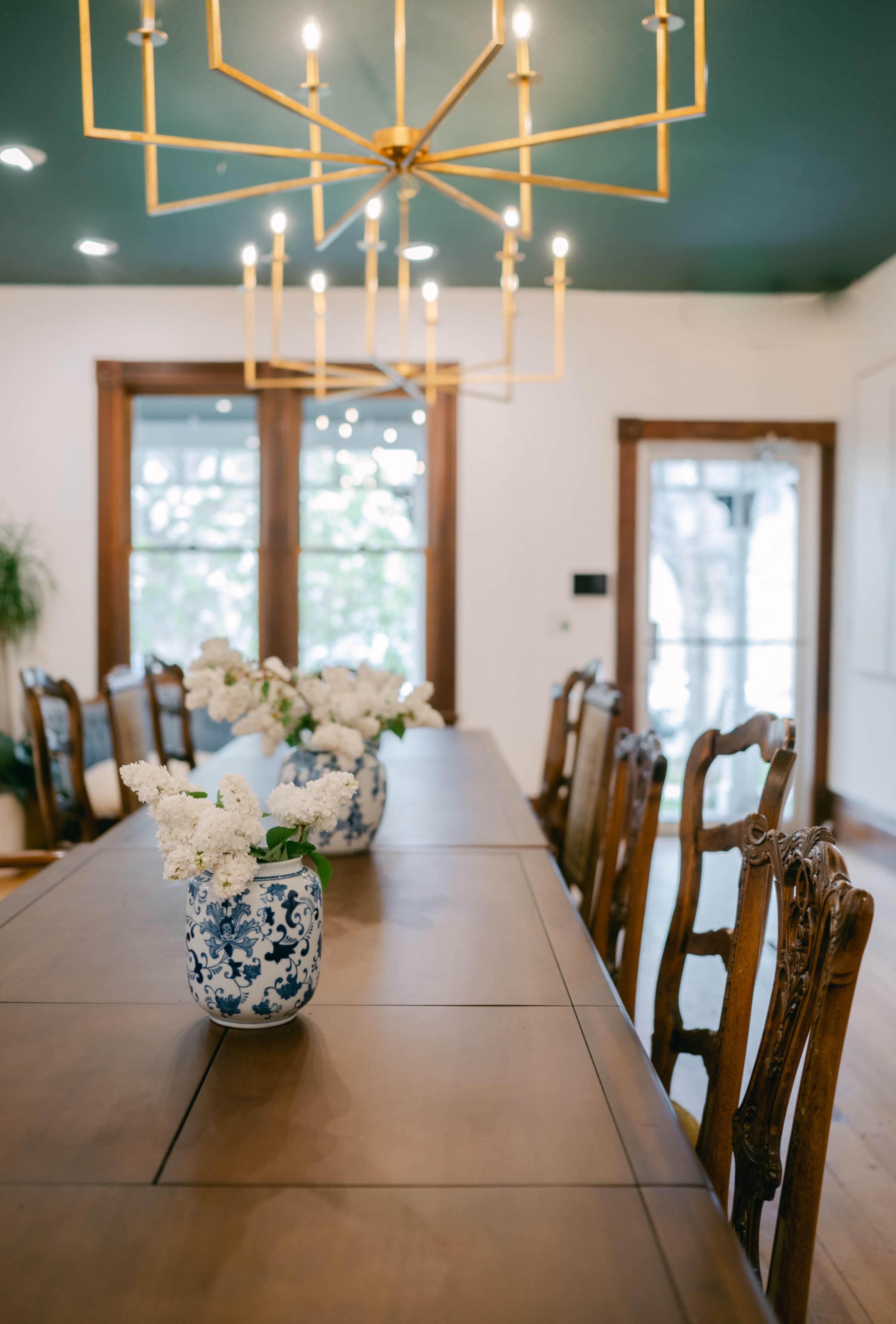 A long wooden dining table, adorned with blue and white vases of flowers, is illuminated by a modern chandelier in a well-lit dining room.