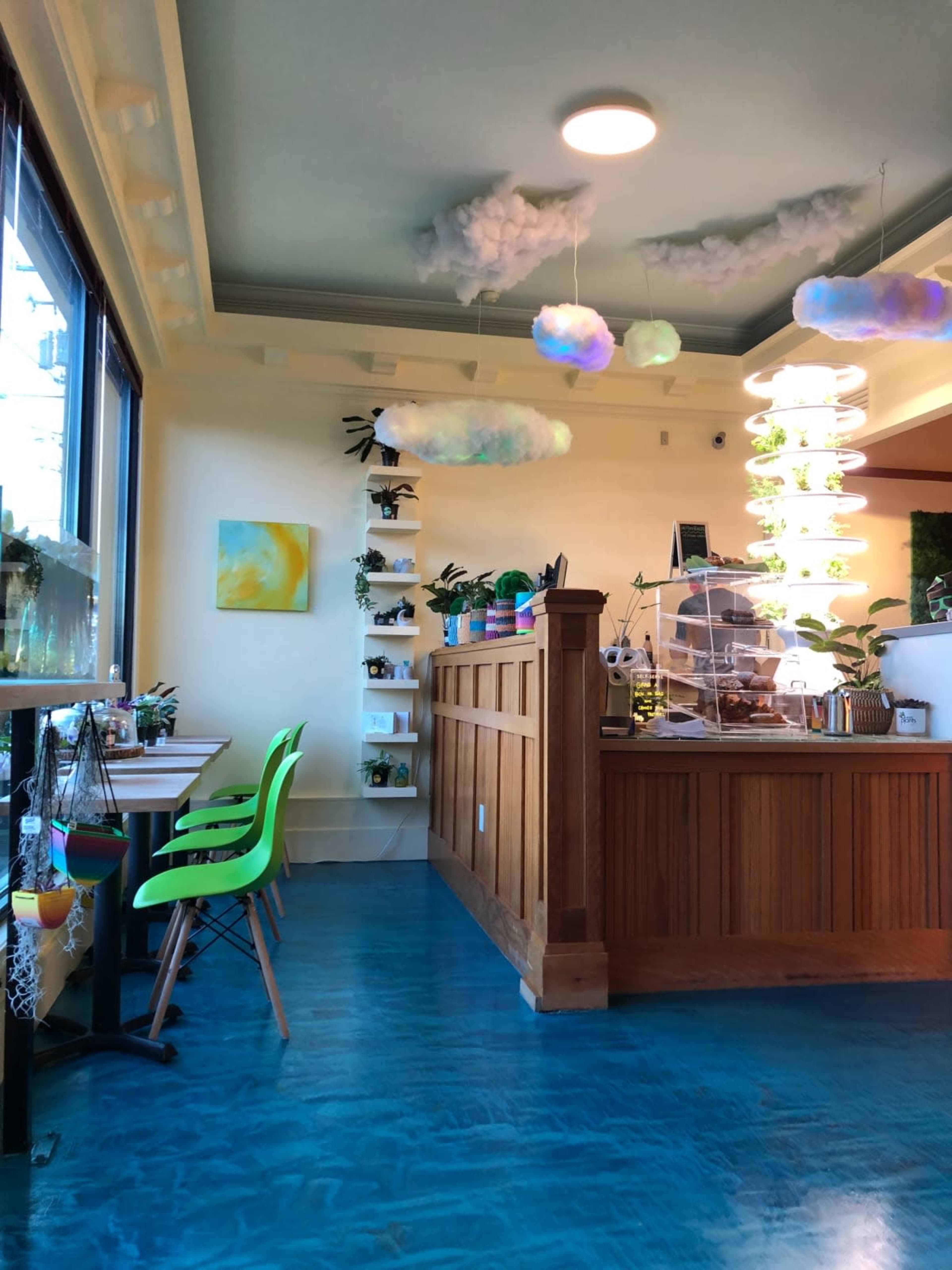 The image shows a brightly lit café interior with green chairs, plants on shelves, and a wooden counter decorated with hanging lights and cloud-shaped décor.