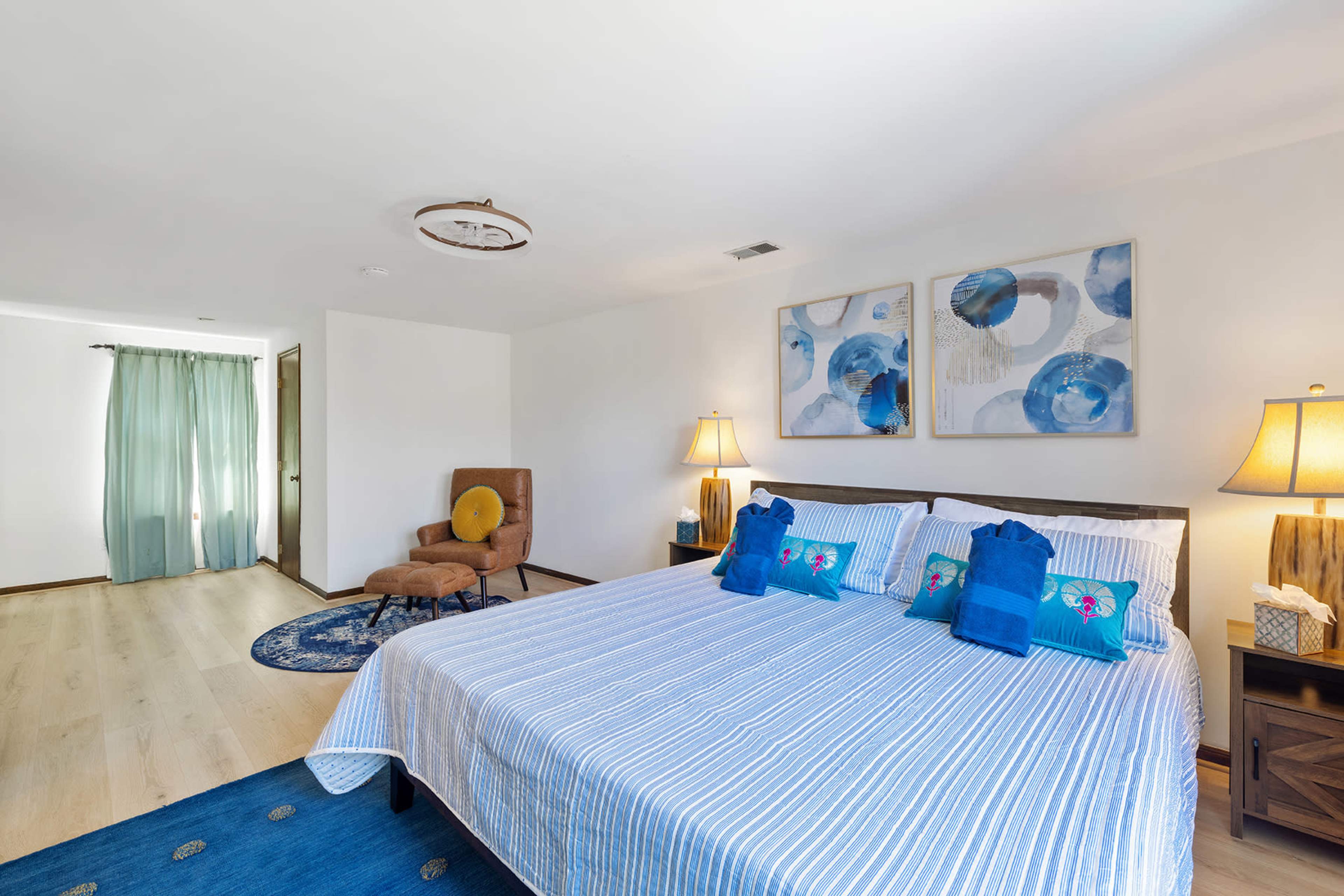 The image shows a bright bedroom featuring a bed with blue and white striped bedding, two lamps on nightstands, and a seating area with a brown chair and decorative blue art on the walls.