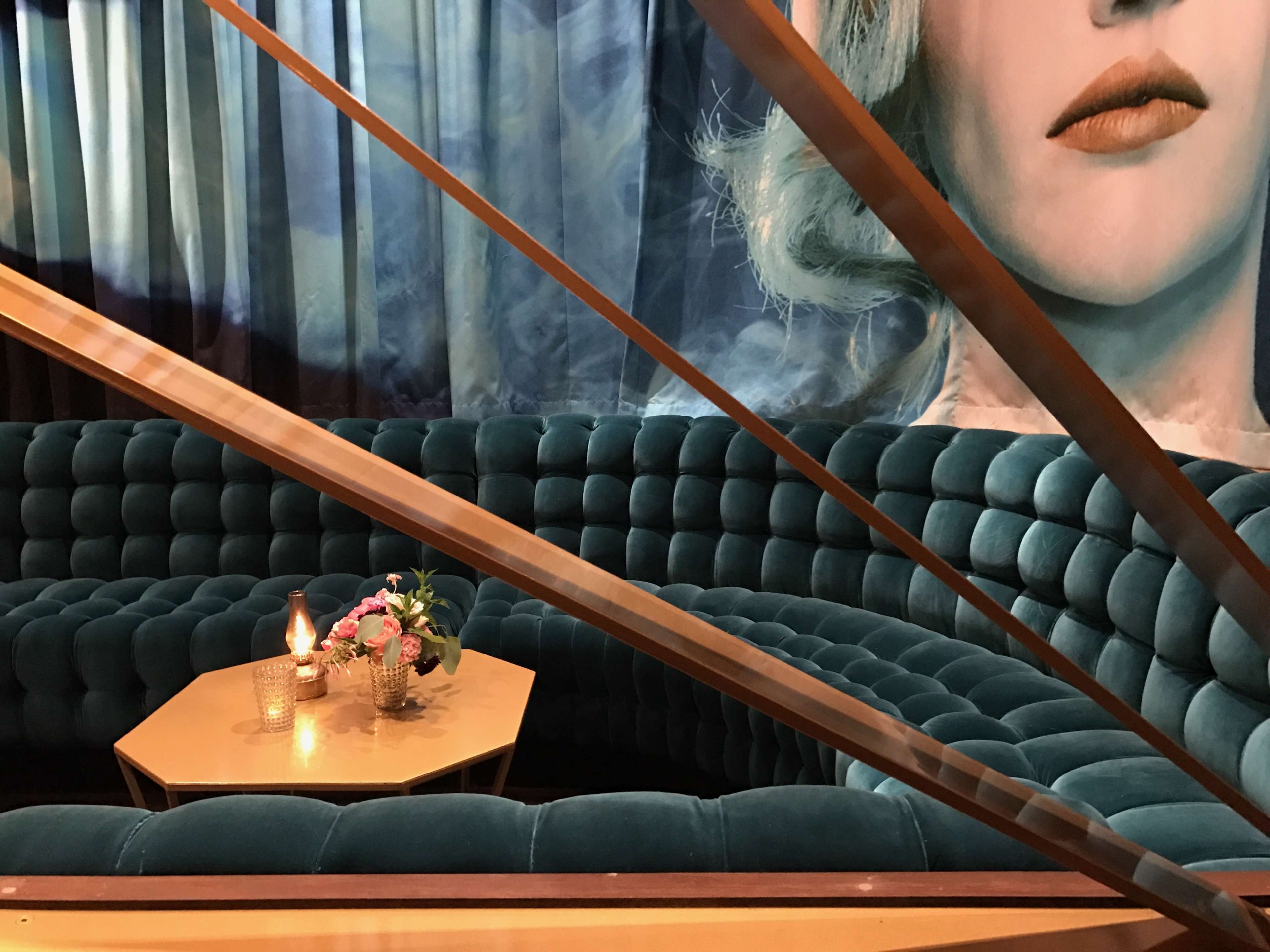 A plush, teal velvet seating area with a small coffee table adorned with flowers and a candle, set against a backdrop featuring a large mural of a woman's face.