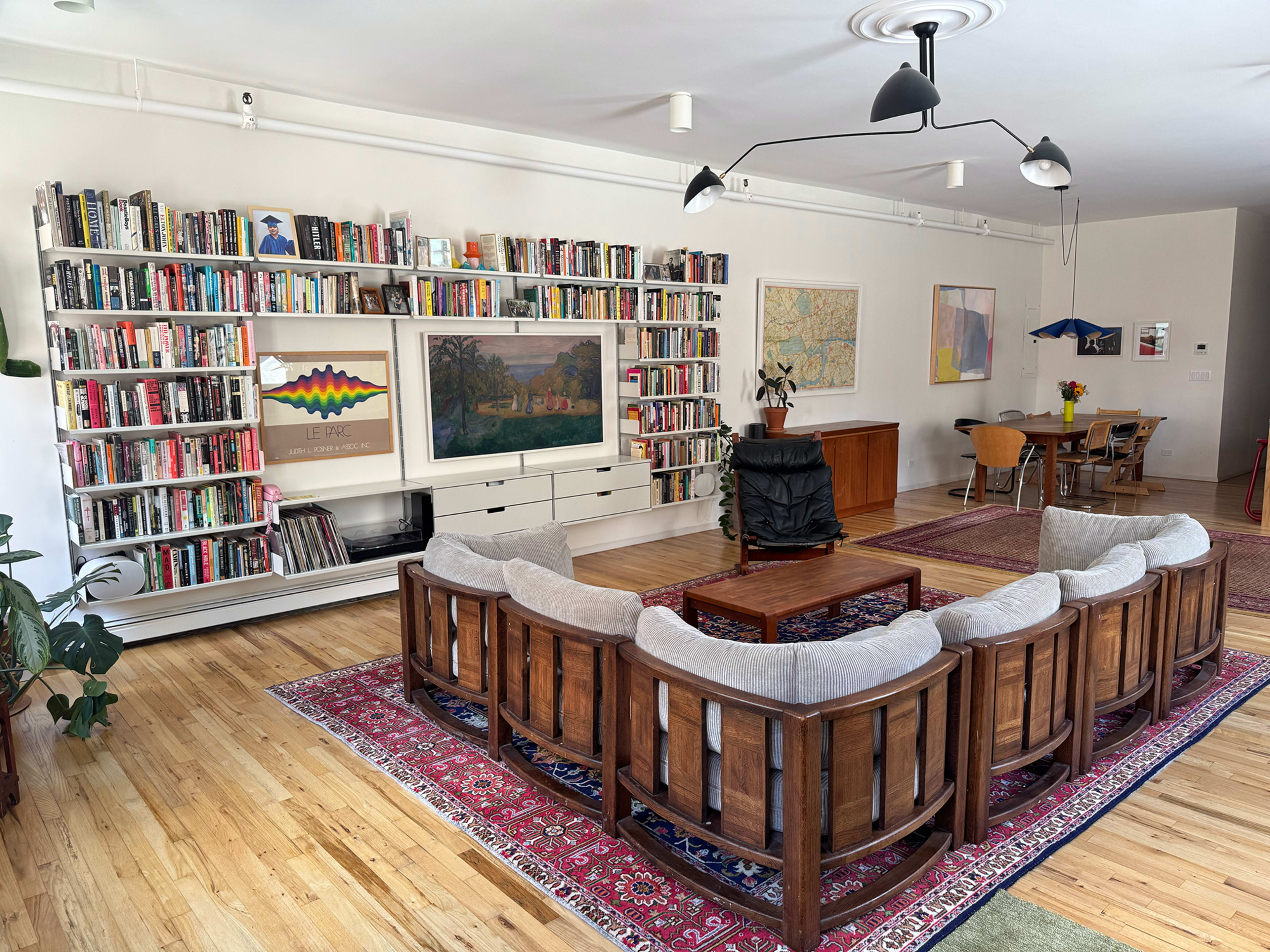The image shows a spacious living room featuring a large bookshelf filled with books, a circular arrangement of wooden chairs, a coffee table in the center, and a dining area in the background.