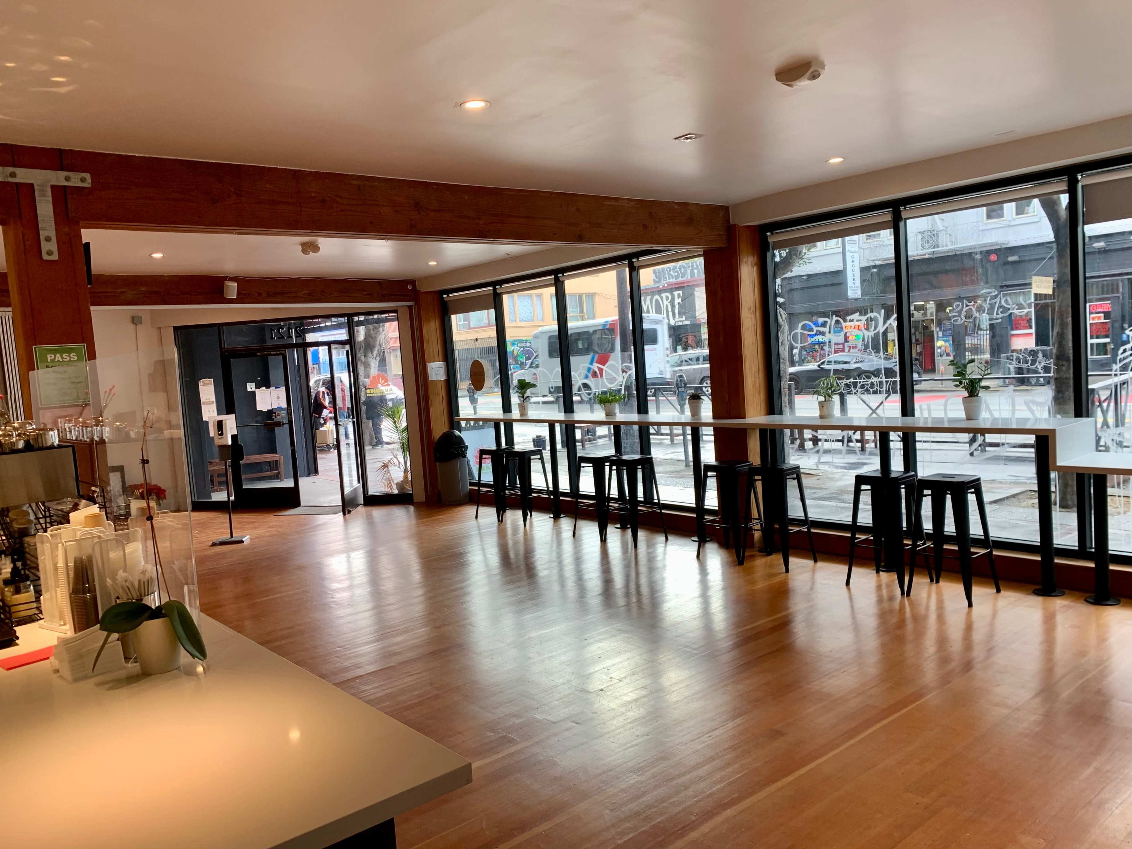 An interior of a cafe with wooden beams, large windows allowing a view of a street with graffiti, and black high stools arranged near a long, white table.