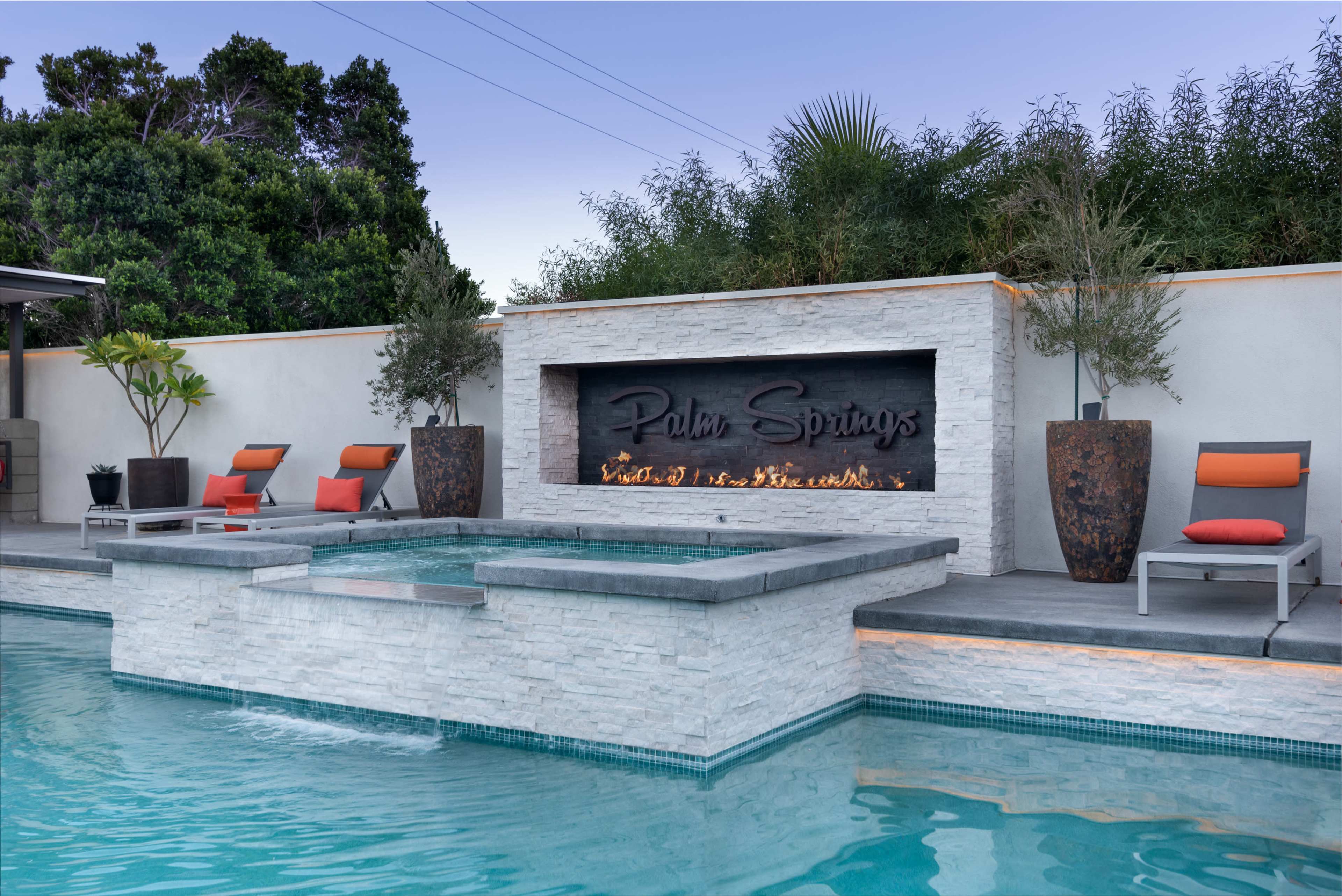 A modern backyard with a swimming pool, spa, and a stone wall featuring the text "Palm Springs," surrounded by minimalist landscaping and patio furniture.