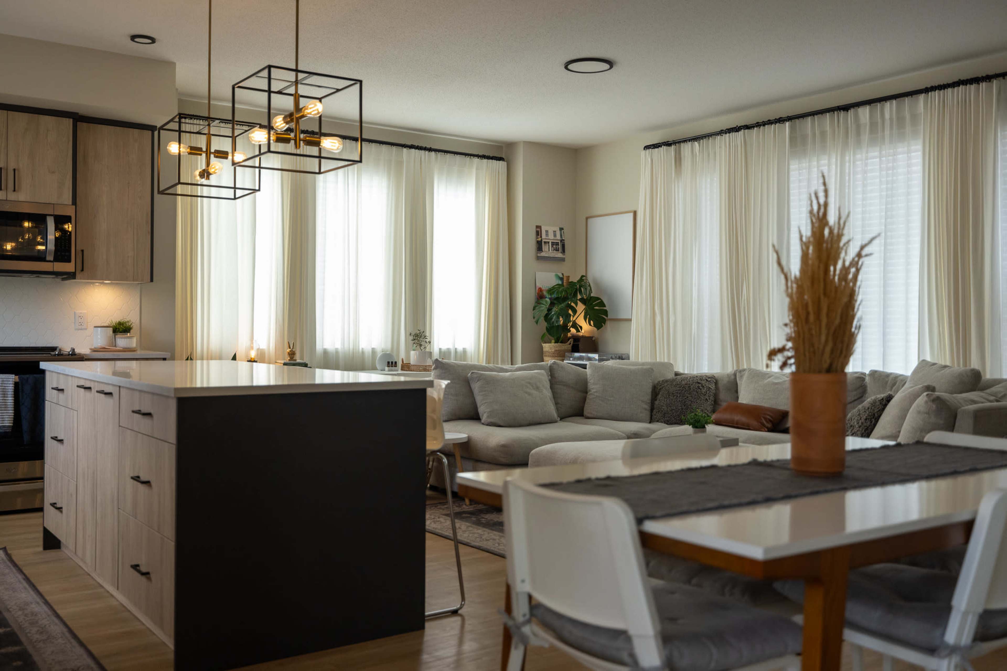 A modern living space featuring a kitchen island, a dining table, comfortable seating, and large windows with sheer curtains.