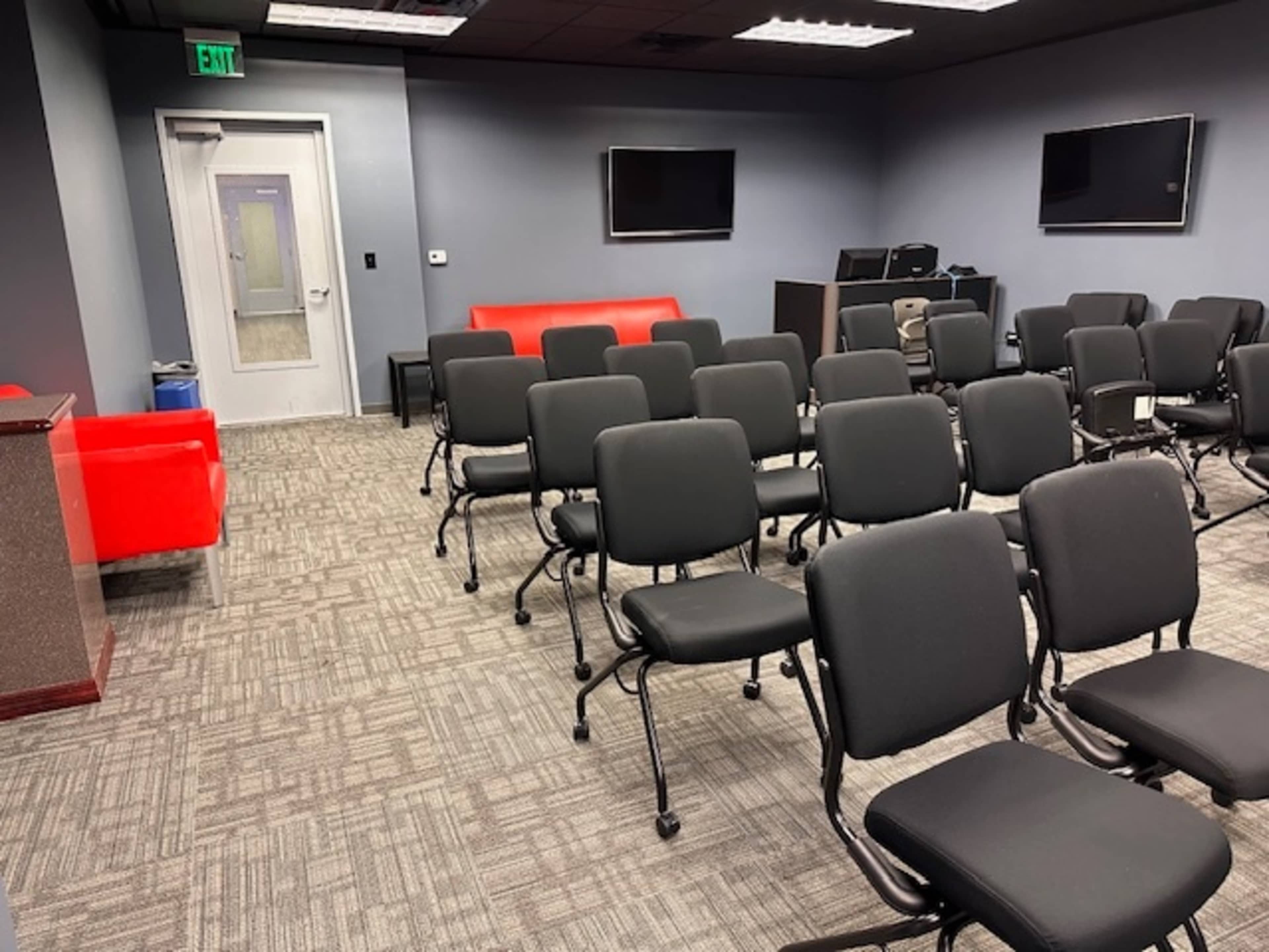 The image shows a conference room with several rows of black chairs facing a red couch against a gray wall, along with two wall-mounted televisions.