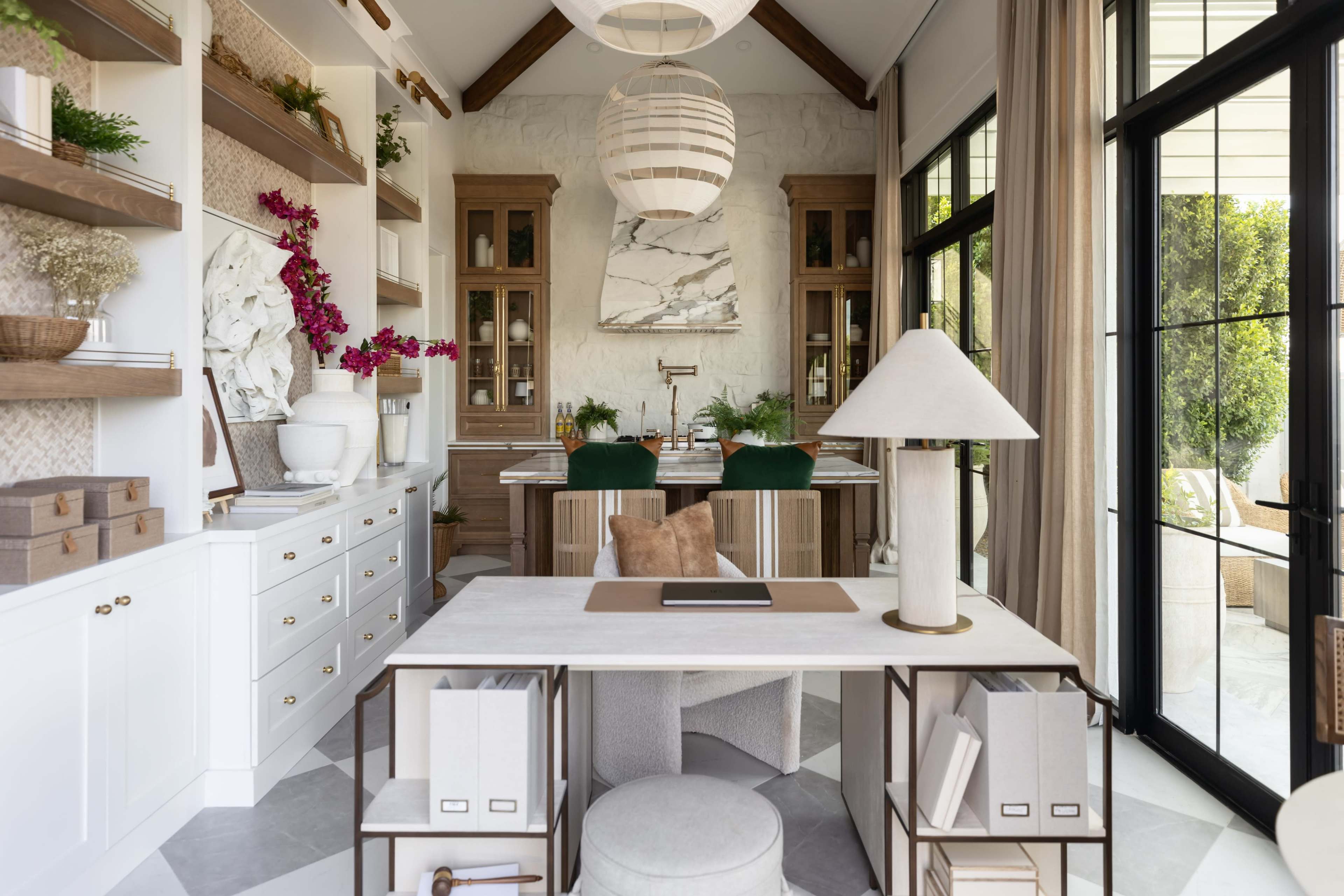 The image shows a modern home office with a large white desk, decorative shelves, and ample natural light from large windows.