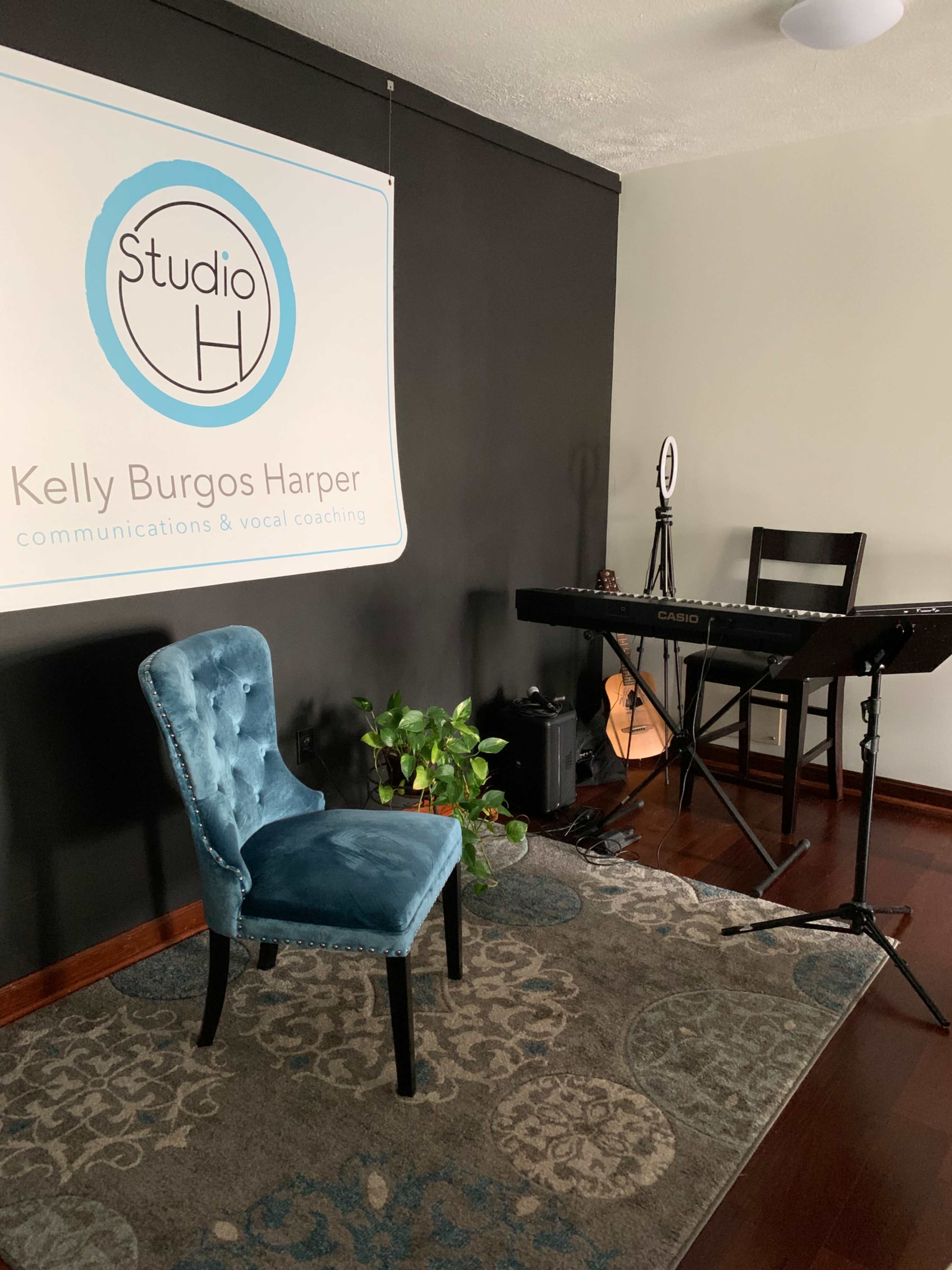 The image shows a vocal coaching studio featuring a blue chair, a keyboard, and a backdrop with the name "Studio H" along with a logo and the name "Kelly Burgos Harper."