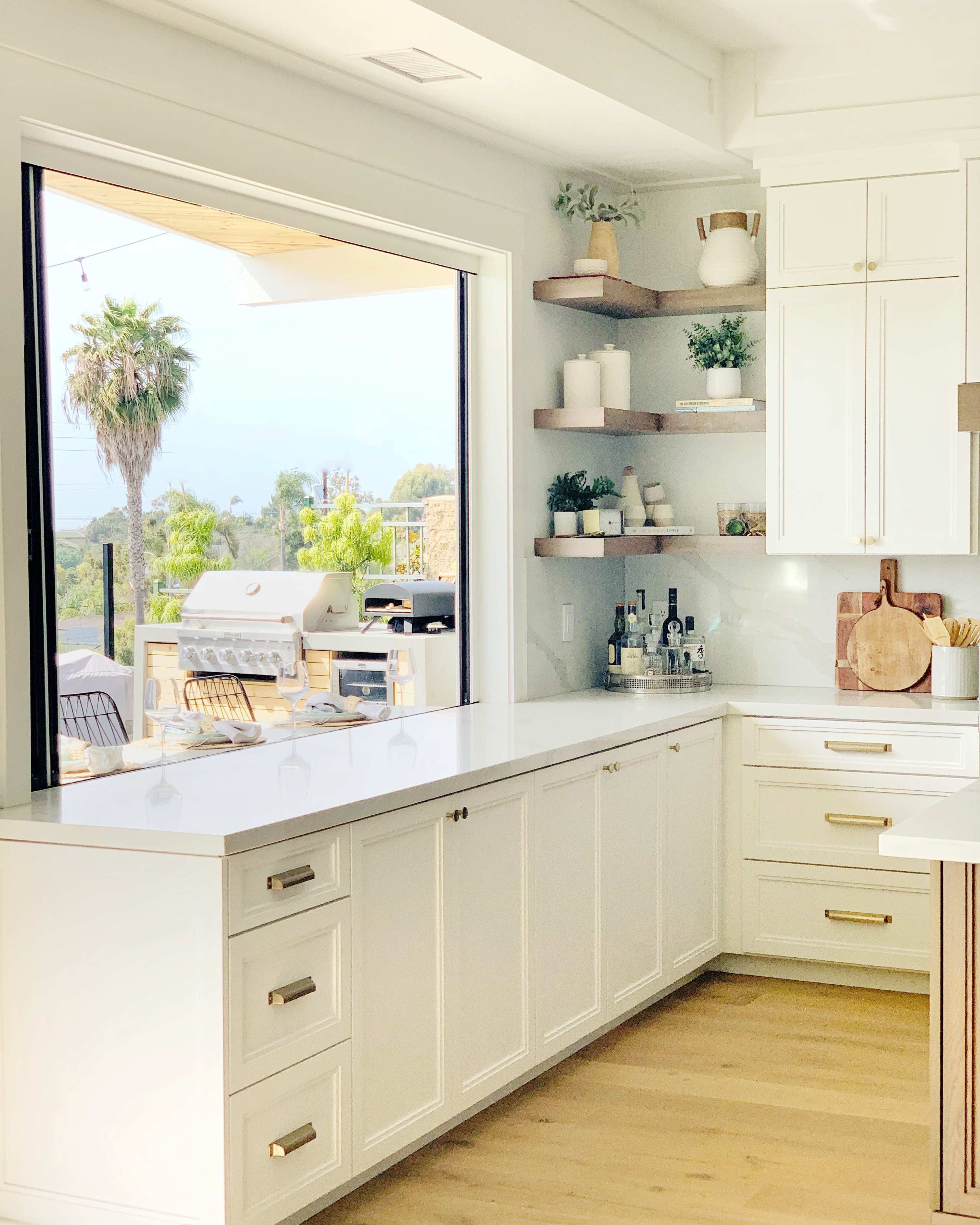A modern kitchen features white cabinetry, a large window with a view of a patio, and wooden shelves displaying plants and barware.