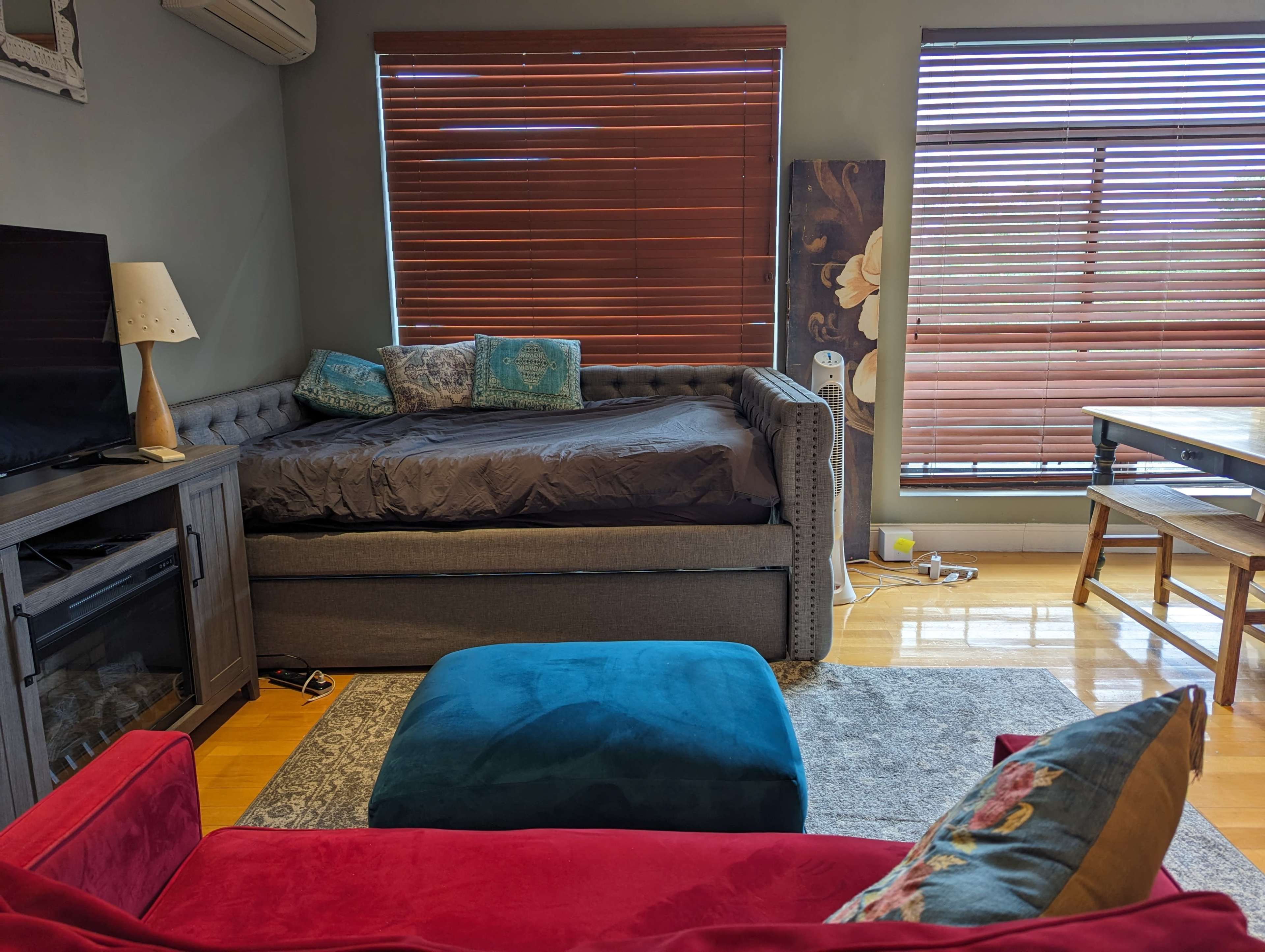 The image shows a cozy living space with a daybed, a television stand, and a dining table, all illuminated by sunlight through wooden blinds.