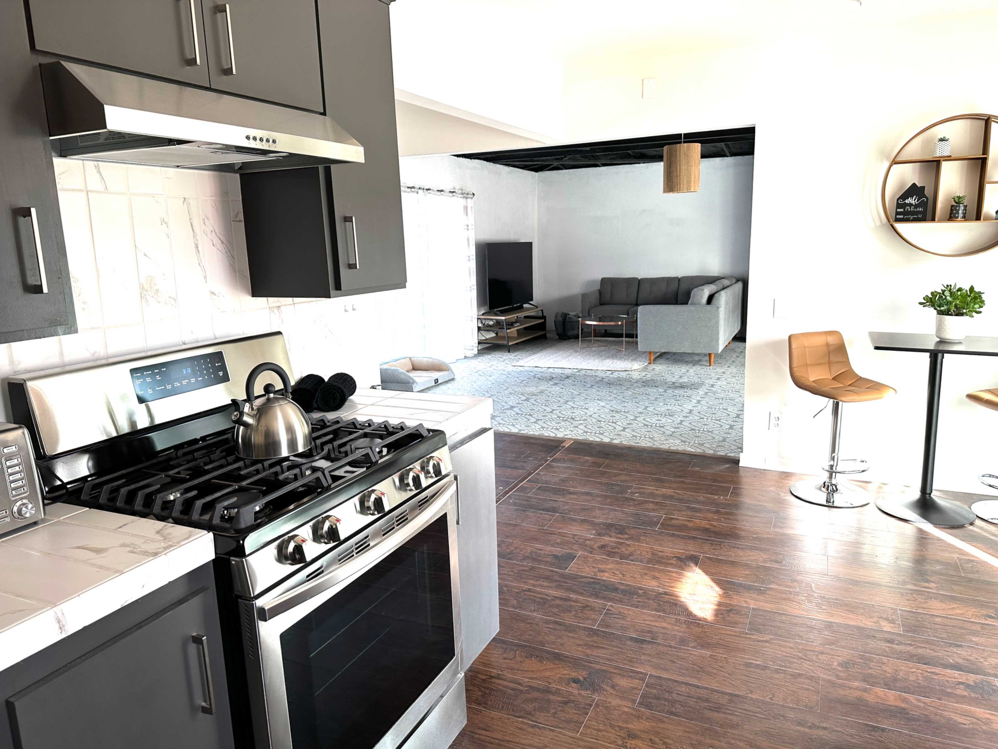 A kitchen with dark cabinetry, stainless steel appliances, and a view of a living area featuring a gray sofa and a small dining set in the background.