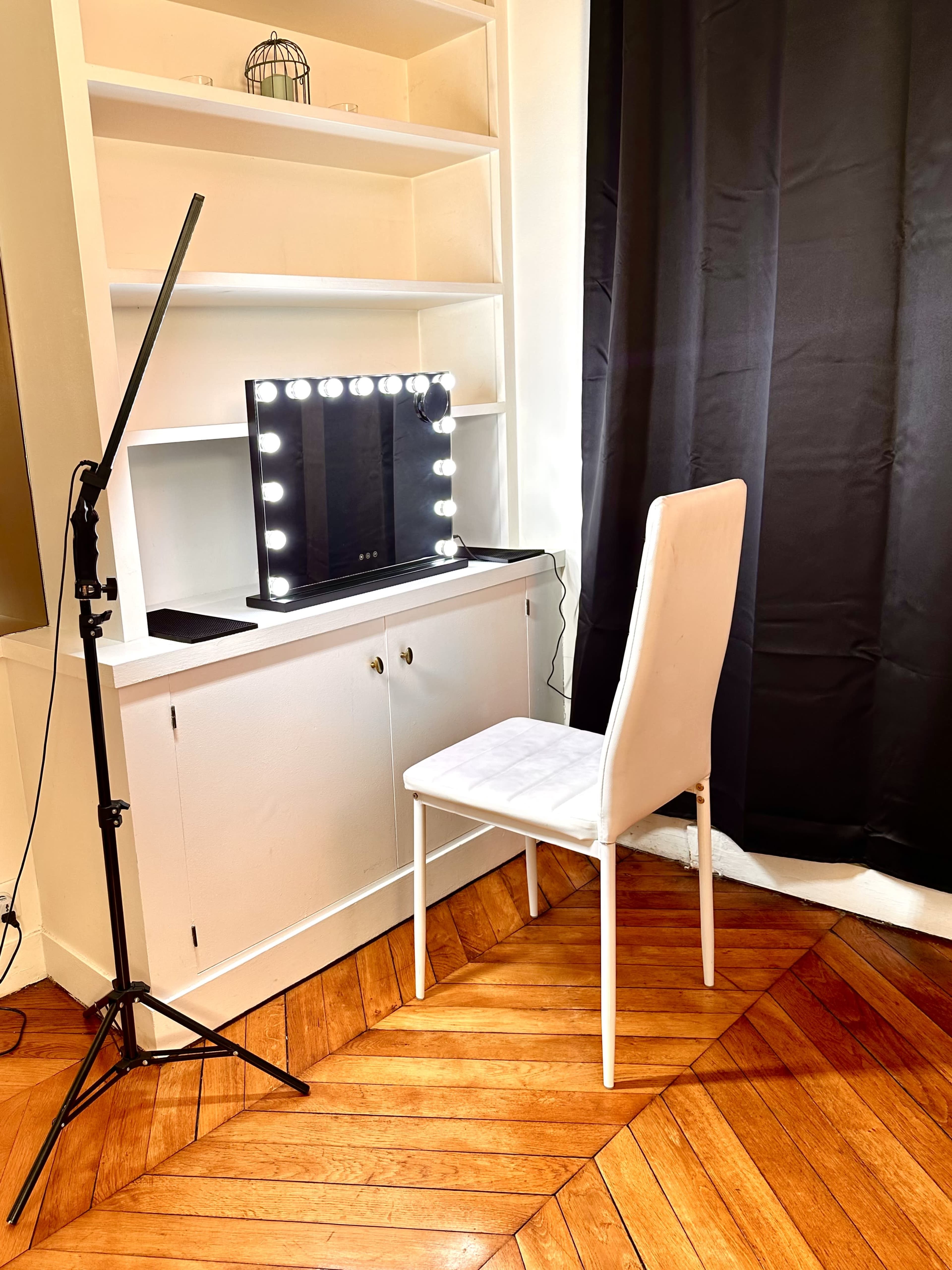 The image shows a makeup vanity with a lit mirror, a white chair, and shelving above it, all positioned against a dark curtain.