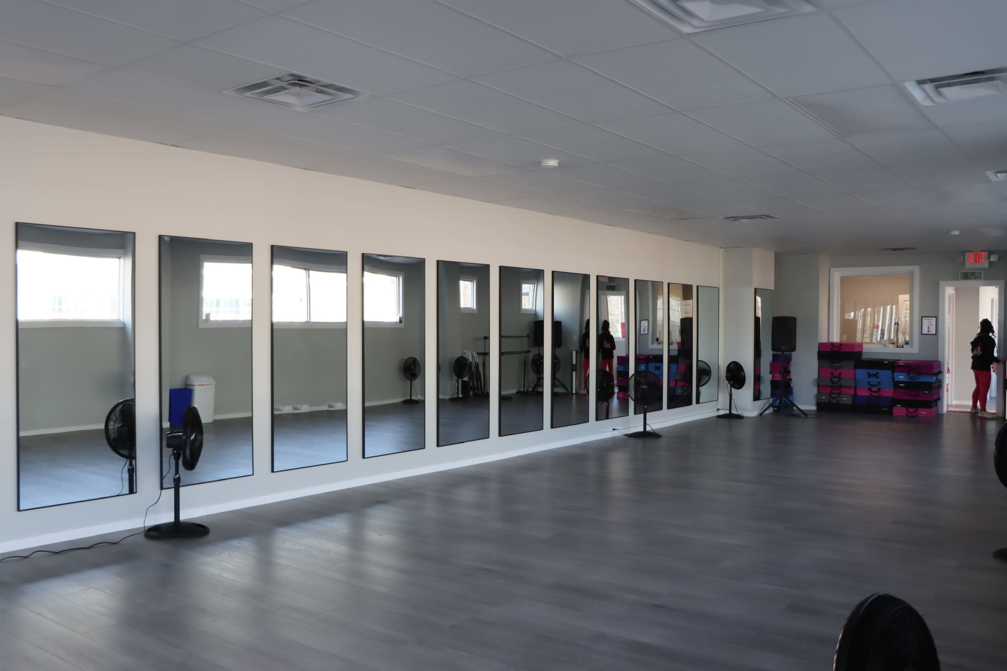 A spacious fitness studio features large mirrors along one wall and several fans positioned throughout the room.