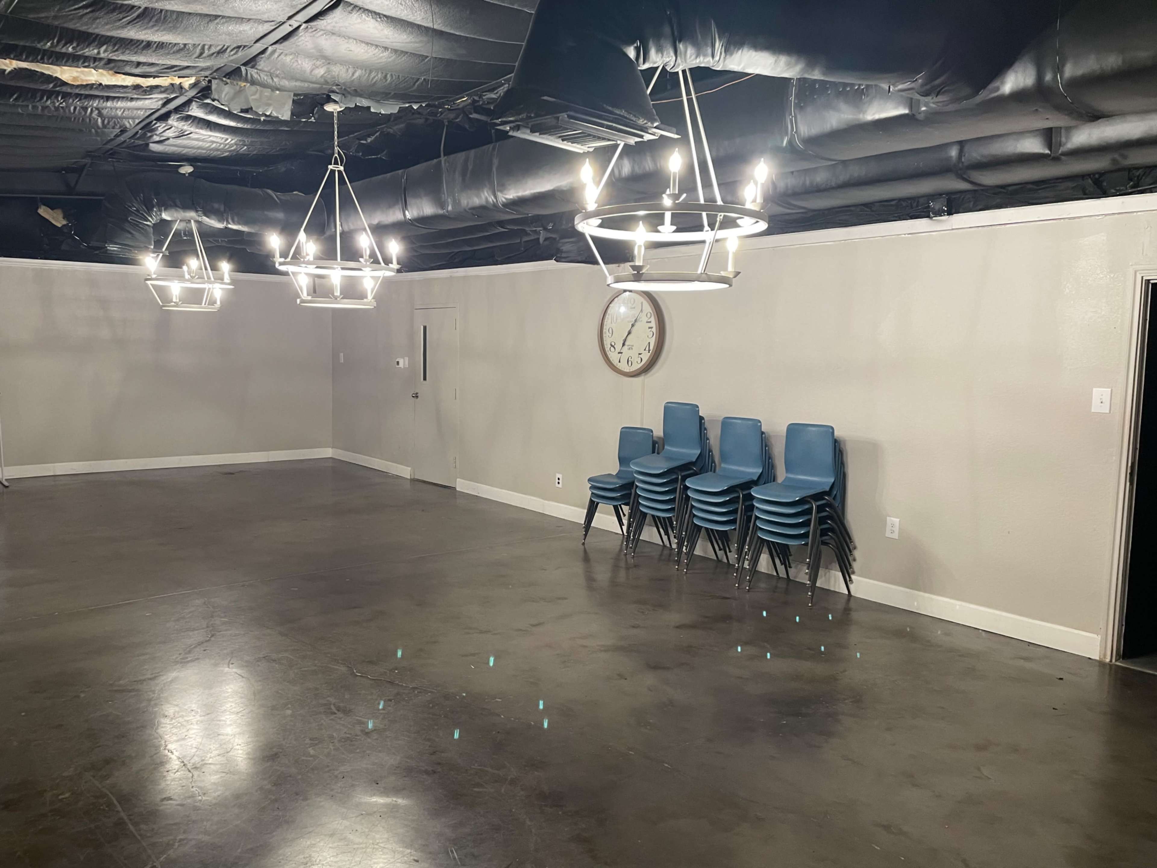 The image shows a vacant room with a stacked set of blue chairs in one corner and modern light fixtures hanging from the ceiling.