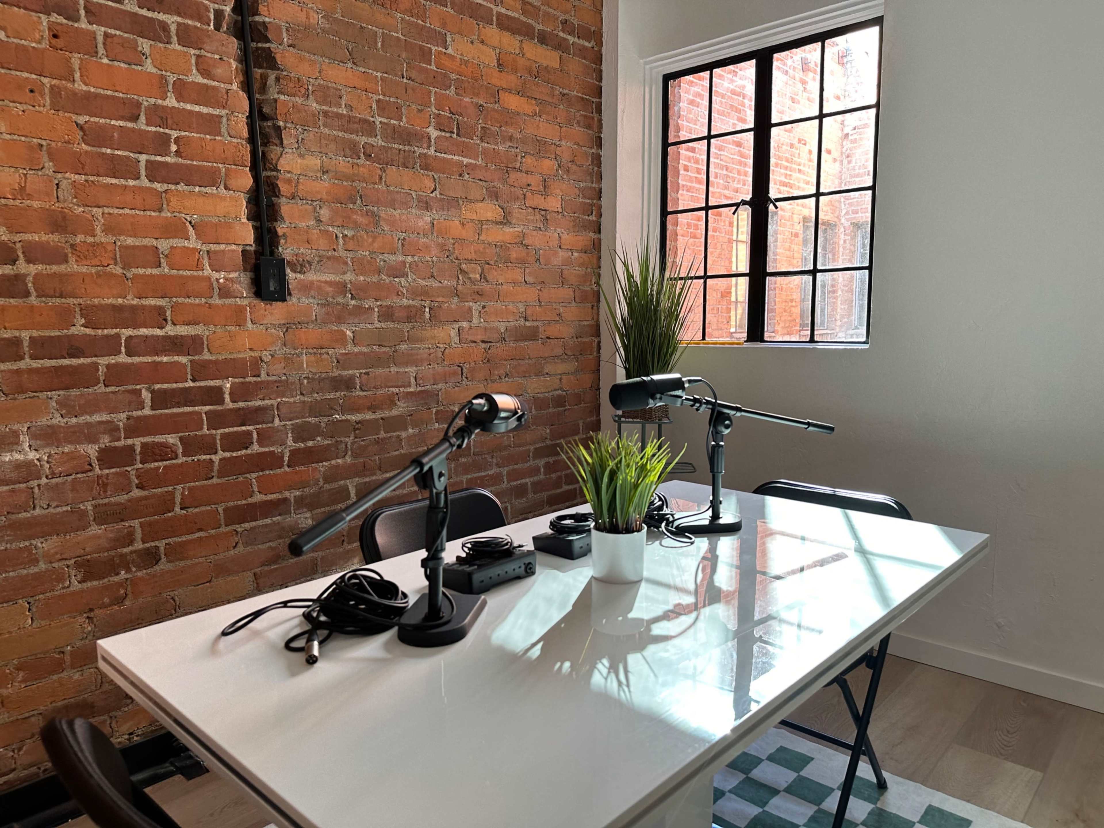 A white table with microphones and cables is set up in a room with brick walls and a window.