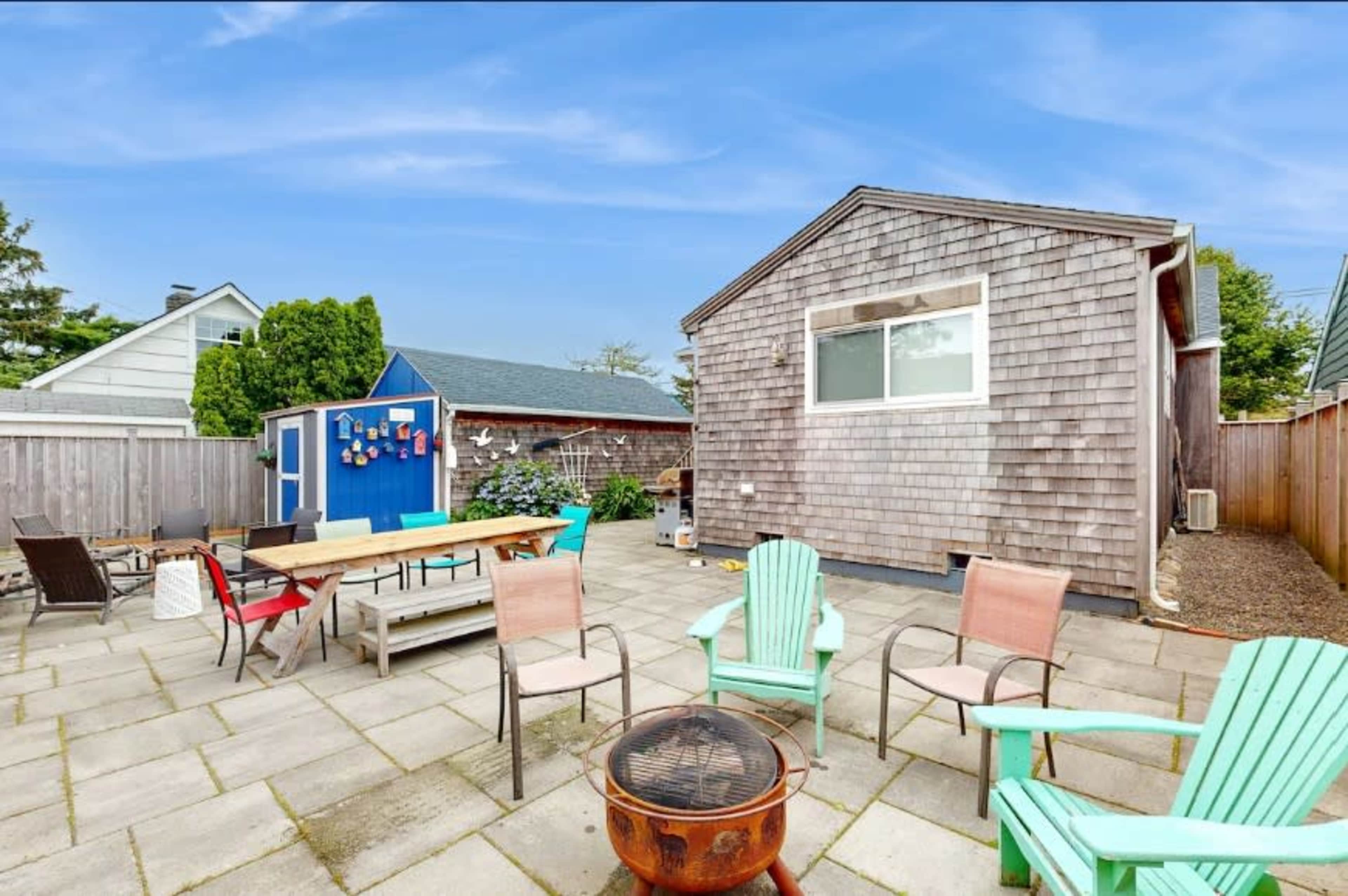 The image shows a backyard patio with a fire pit, surrounded by several chairs and a large wooden table, with a shed and trees in the background.
