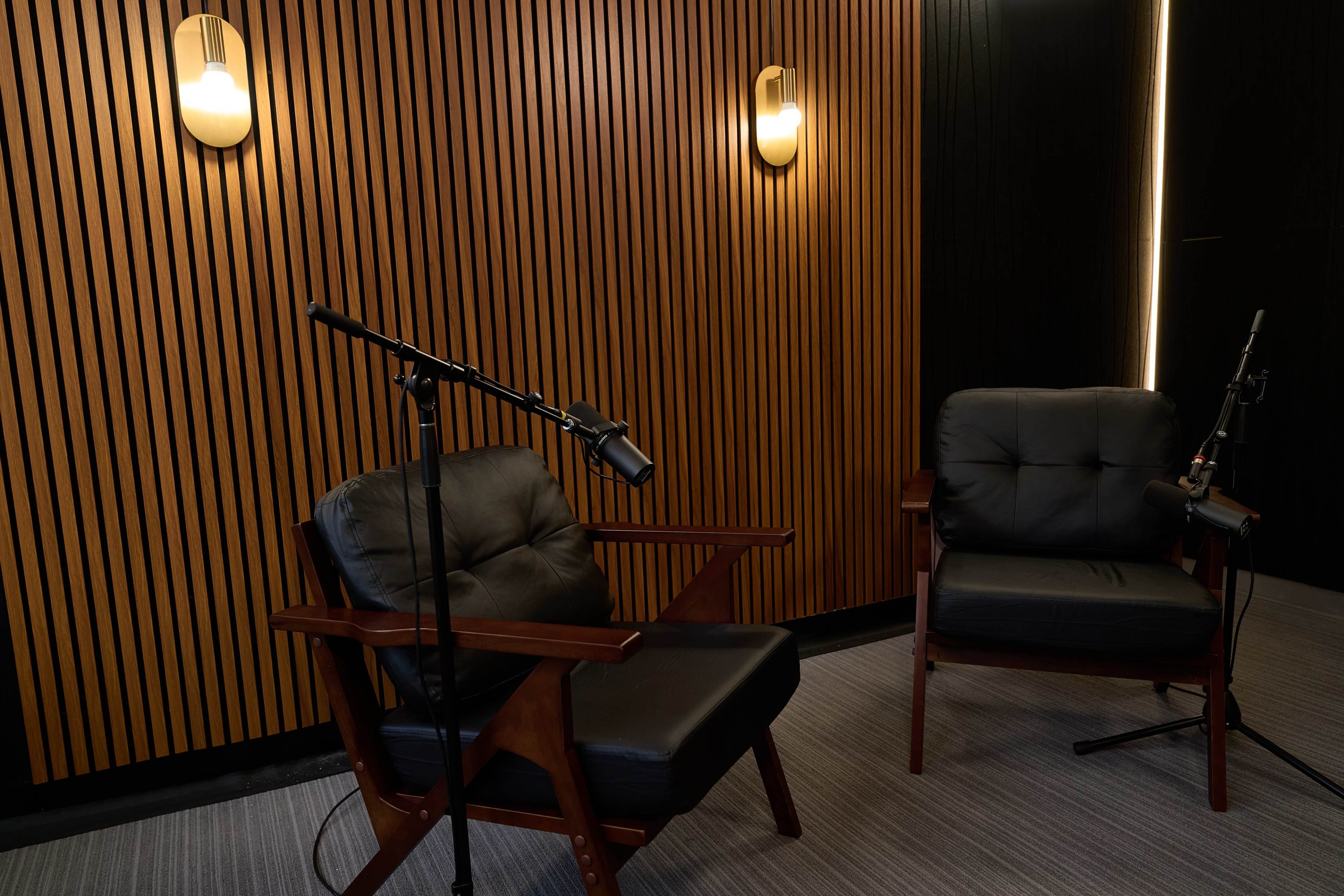The image shows a cozy interview setup featuring two black leather chairs with microphone stands positioned nearby, against a background of wooden slat paneling.
