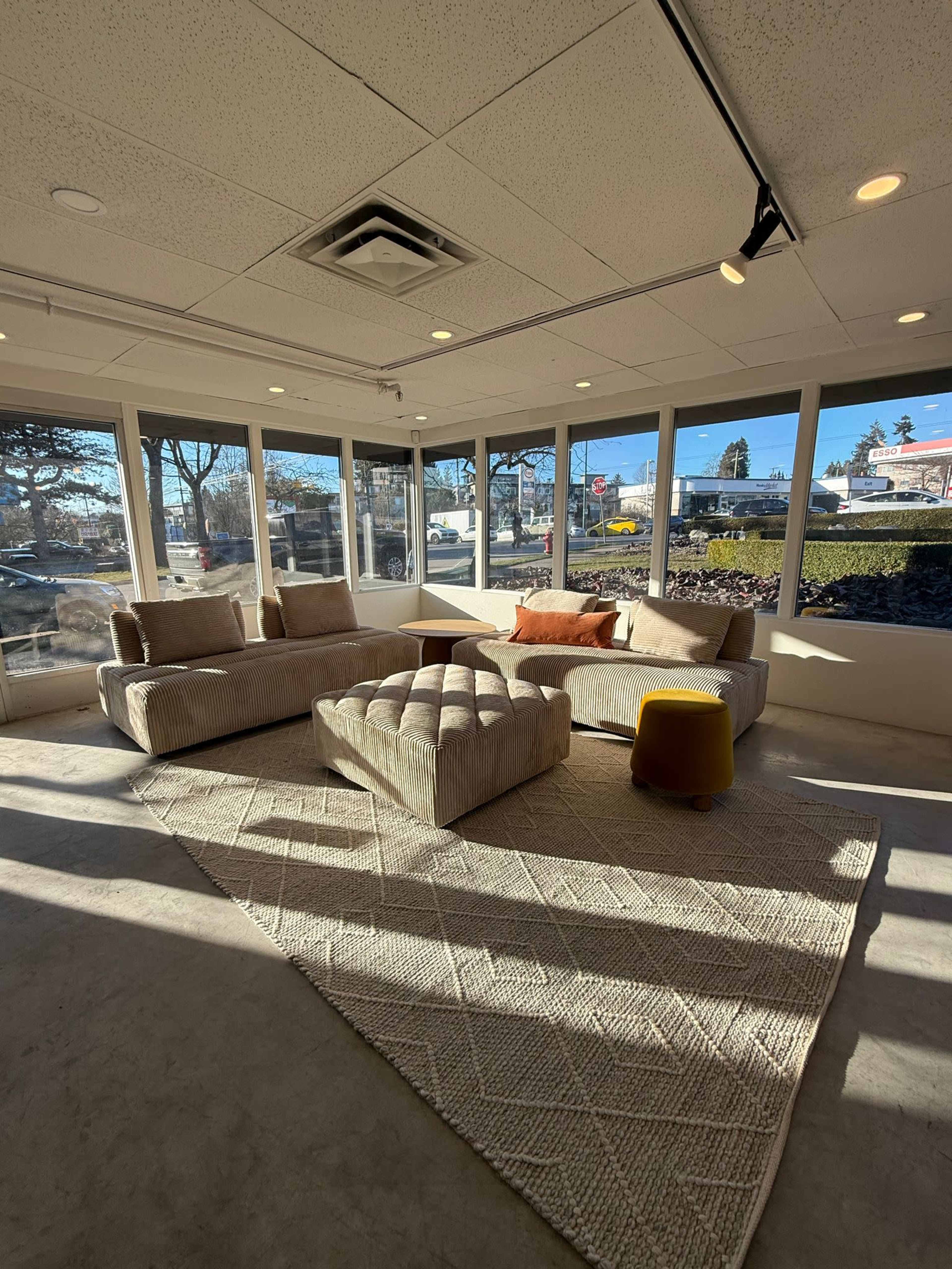 The image shows a modern lounge area with large windows, featuring a sectional sofa, a tufted ottoman, and a round yellow stool on a textured rug.