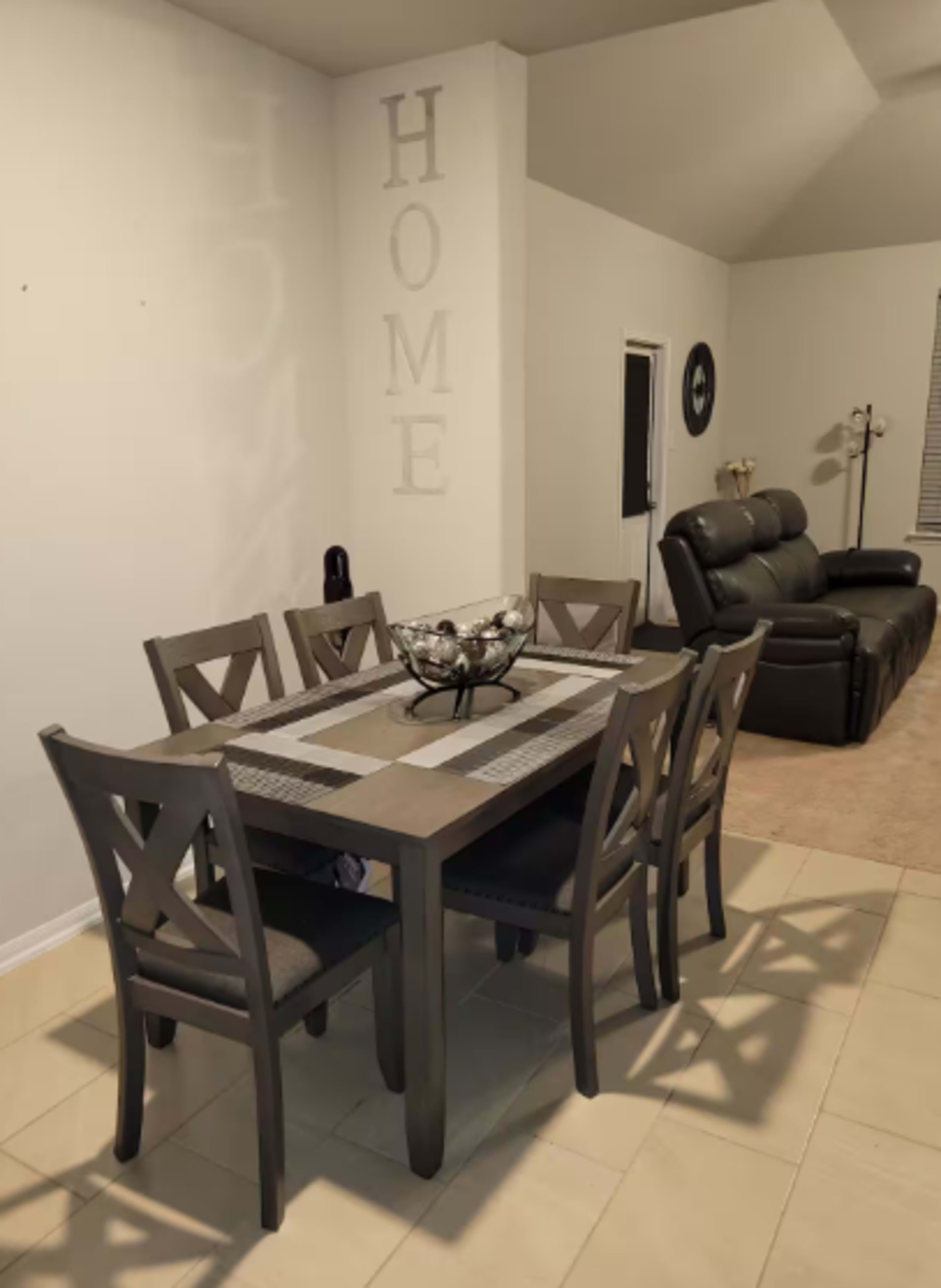 A dining table with six chairs is set against a wall displaying the word "HOME," while a leather sofa sits in the background.