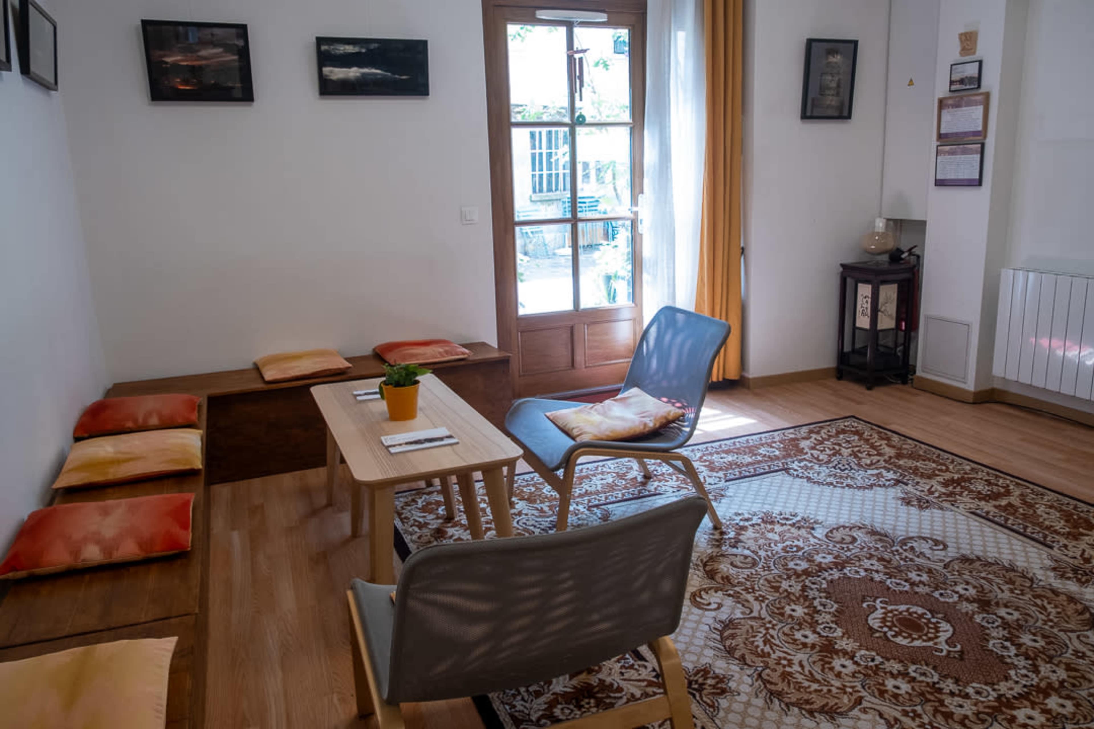 The image shows a cozy room with wooden furniture, a table, seating cushions along the wall, and a large window with soft curtains.