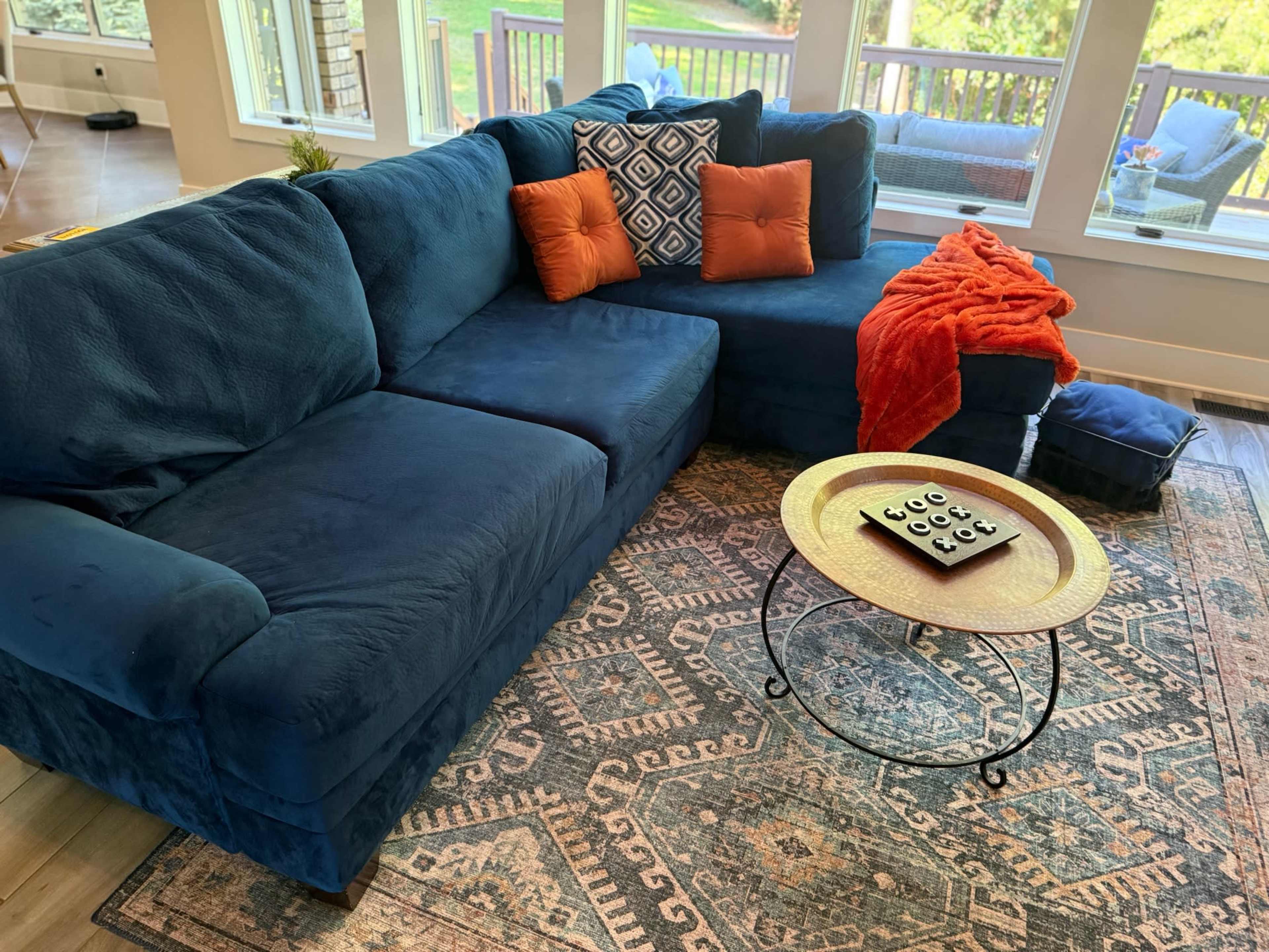The image shows a blue sectional sofa with orange and patterned throw pillows, a blanket draped over the side, a round coffee table with coasters, and a patterned area rug in a well-lit living room.