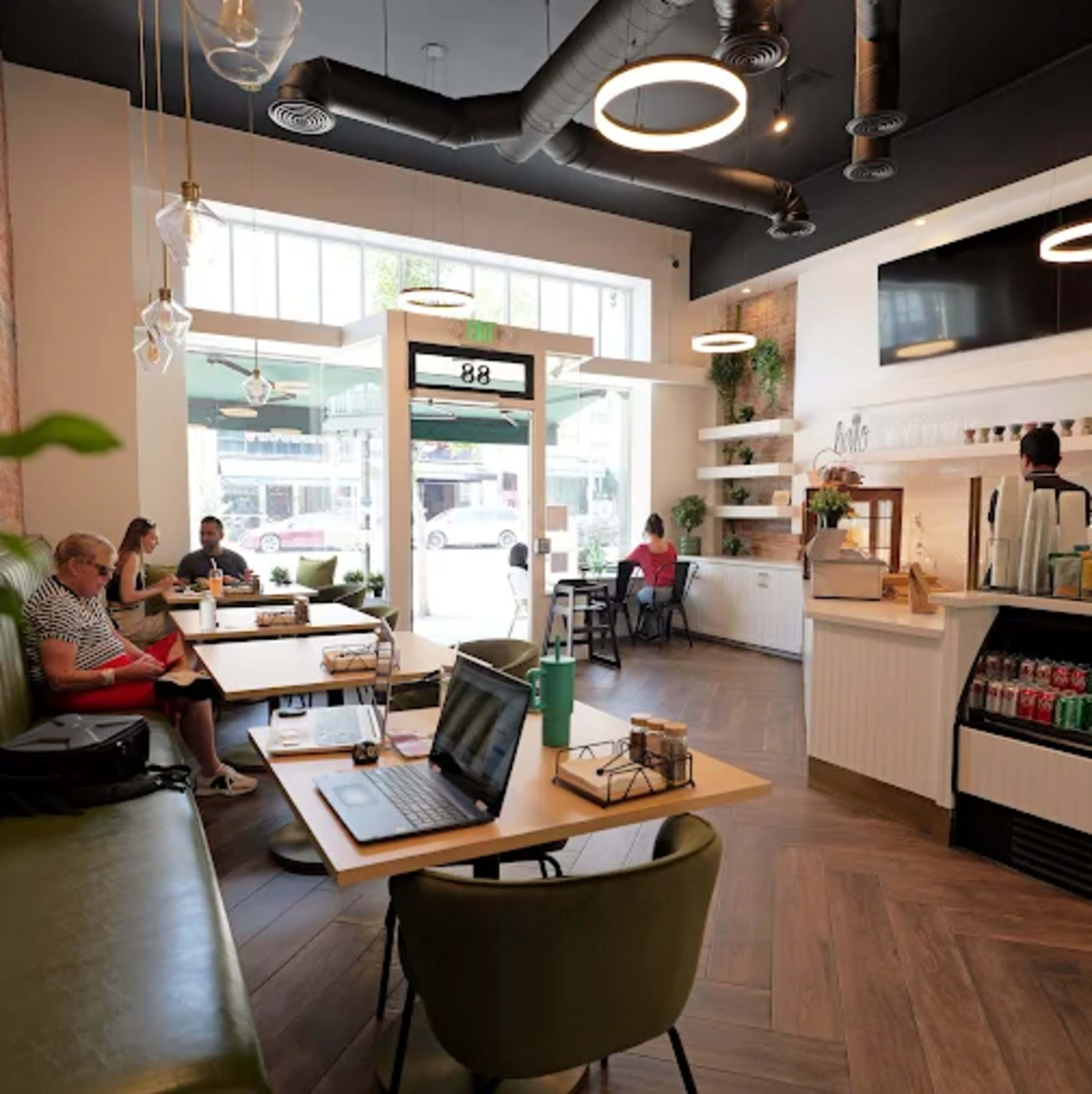 The image shows a modern café interior with green seating, wooden tables, and natural light streaming in from large windows, while customers are engaged in various activities.