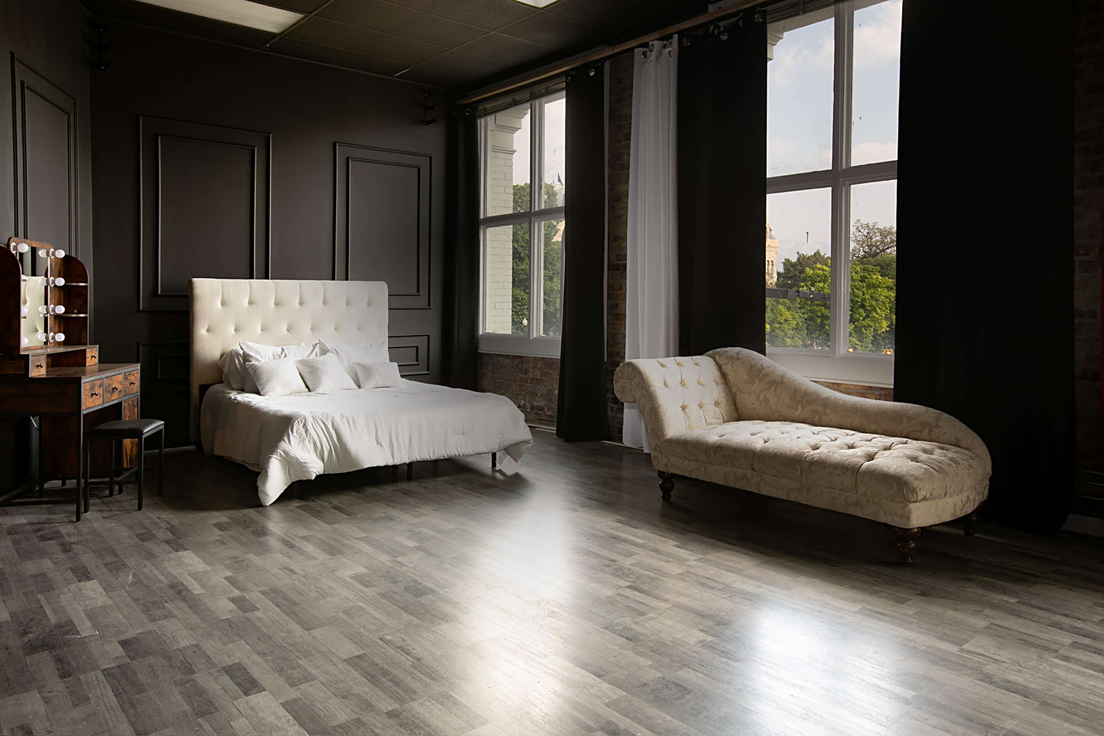 A modern bedroom with a white tufted bed, a vintage chaise lounge, a wooden dresser with a mirror, and large windows letting in natural light.