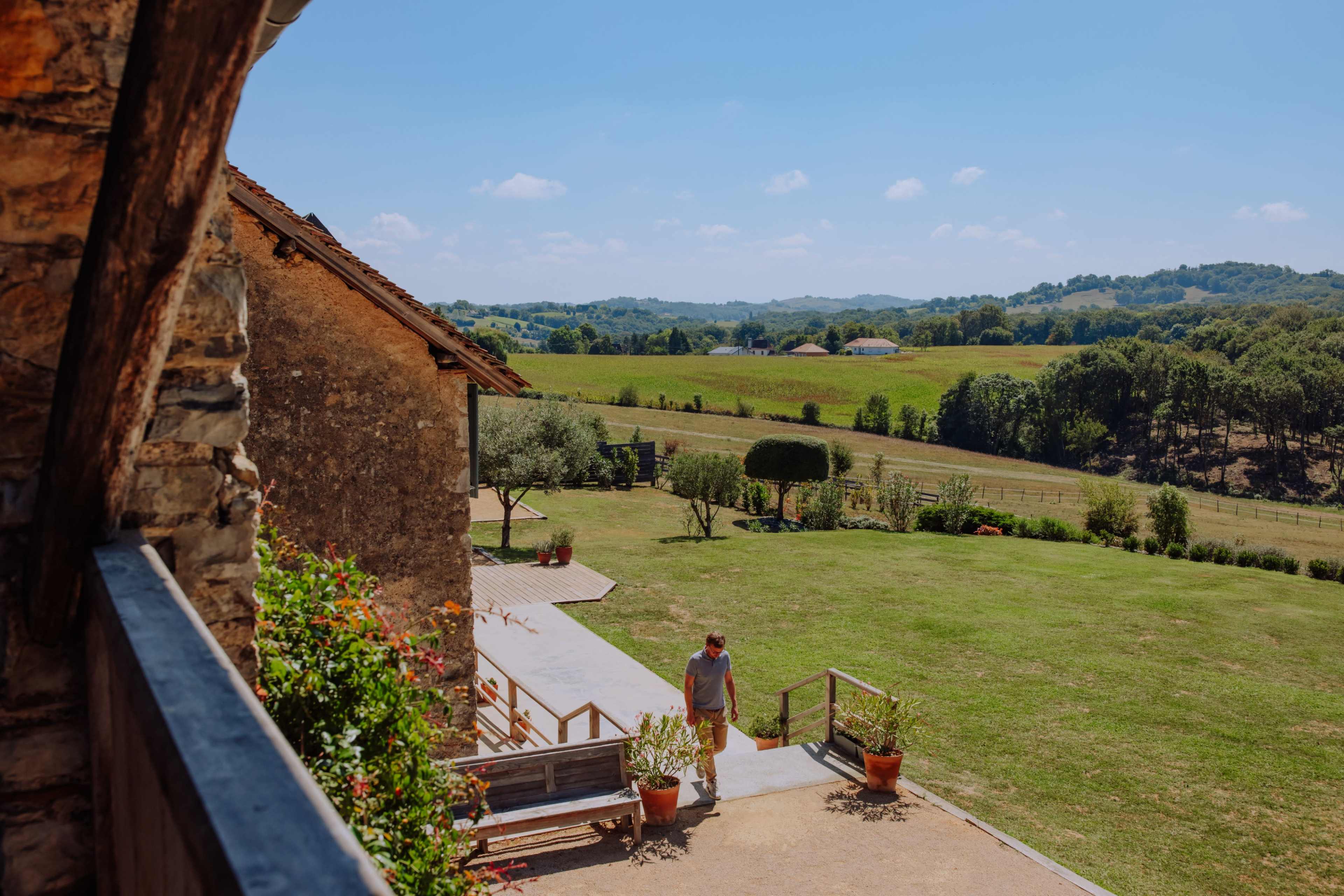 A person walks along a pathway beside a rustic stone building, overlooking a green landscape under a clear blue sky.