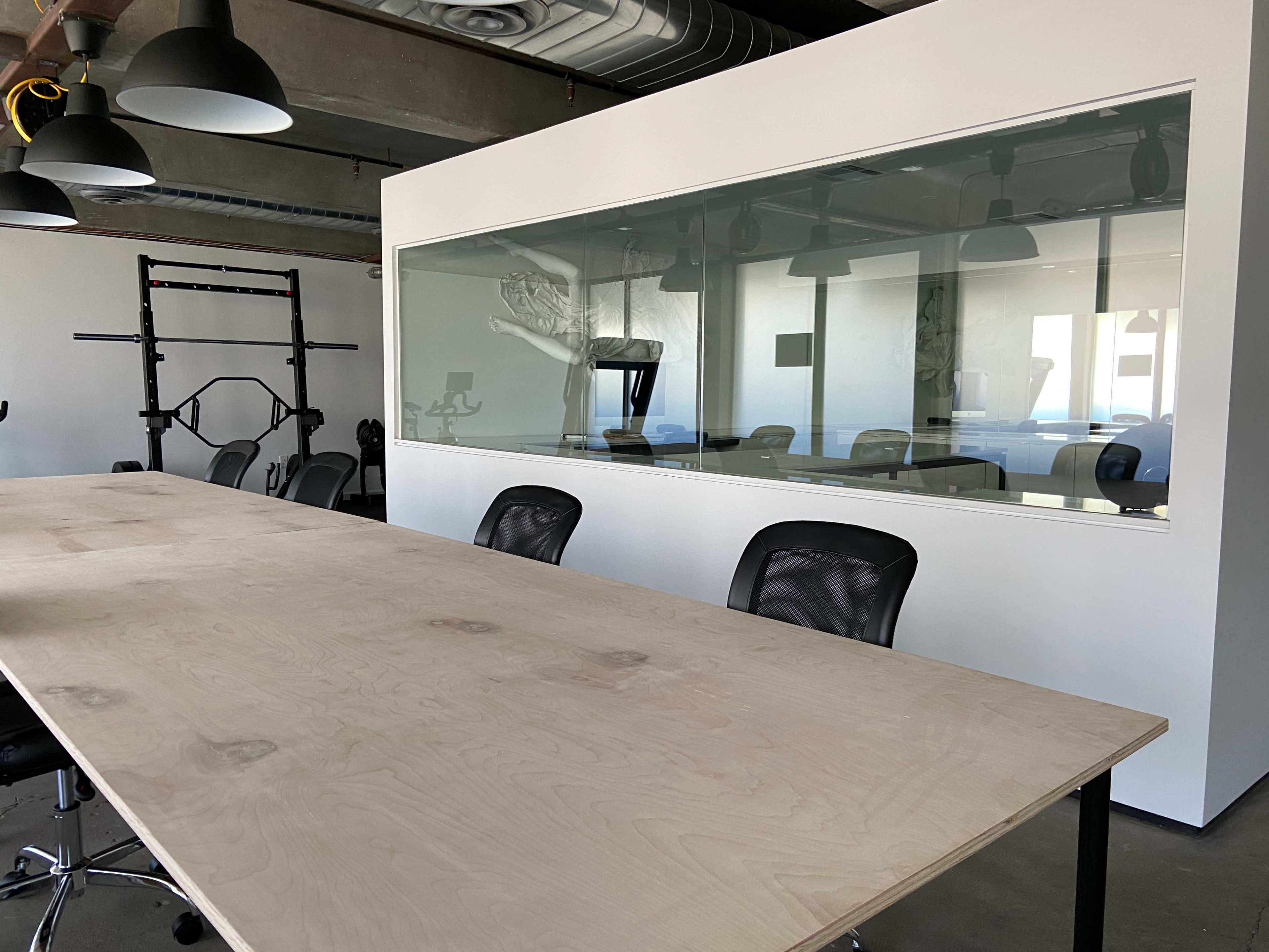 A large wooden conference table with several black chairs is positioned in an office space adjacent to a glass-walled meeting room.