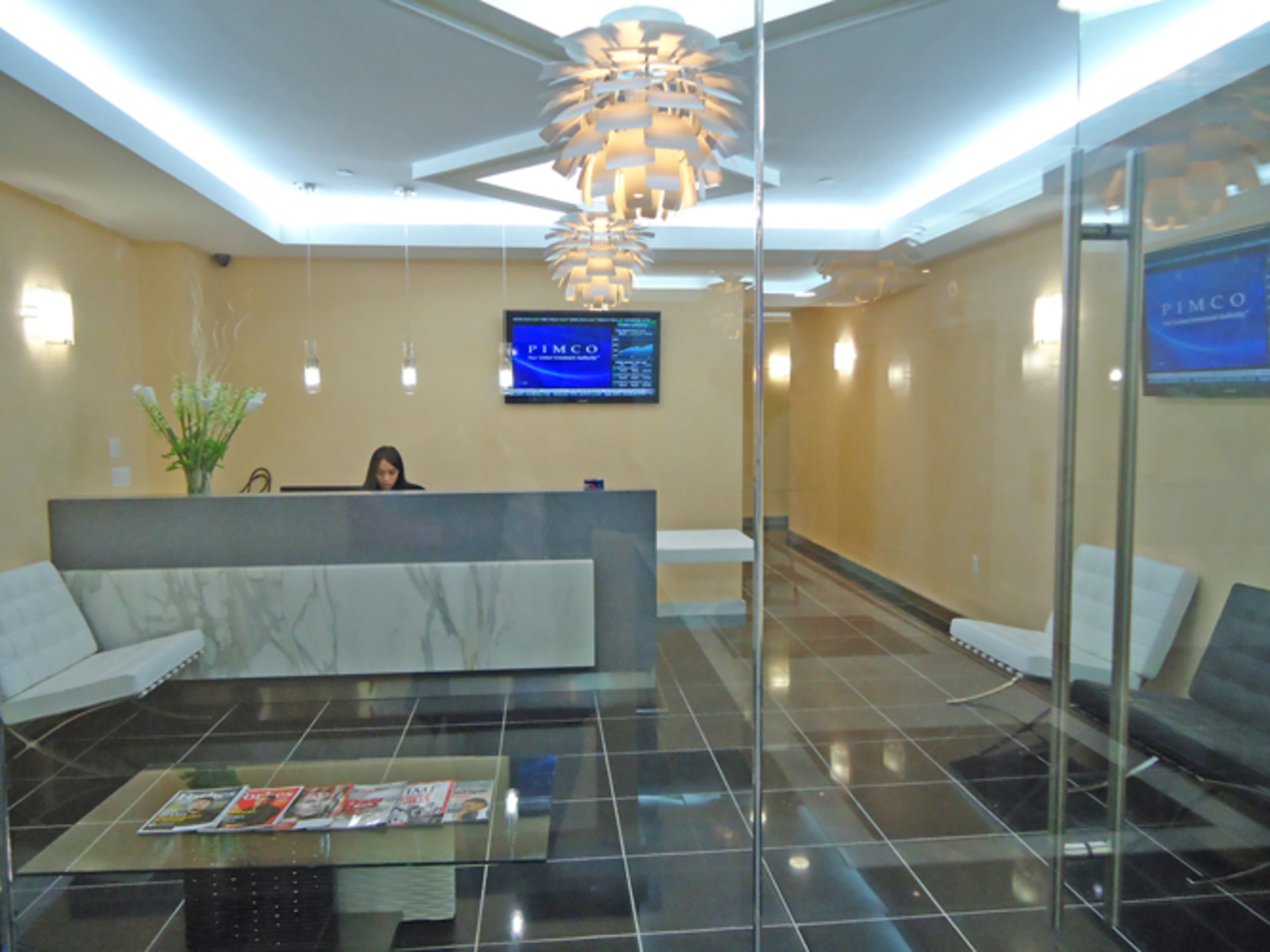 The image shows a modern office reception area with a woman working at a sleek front desk, surrounded by glass walls and minimalist furniture.