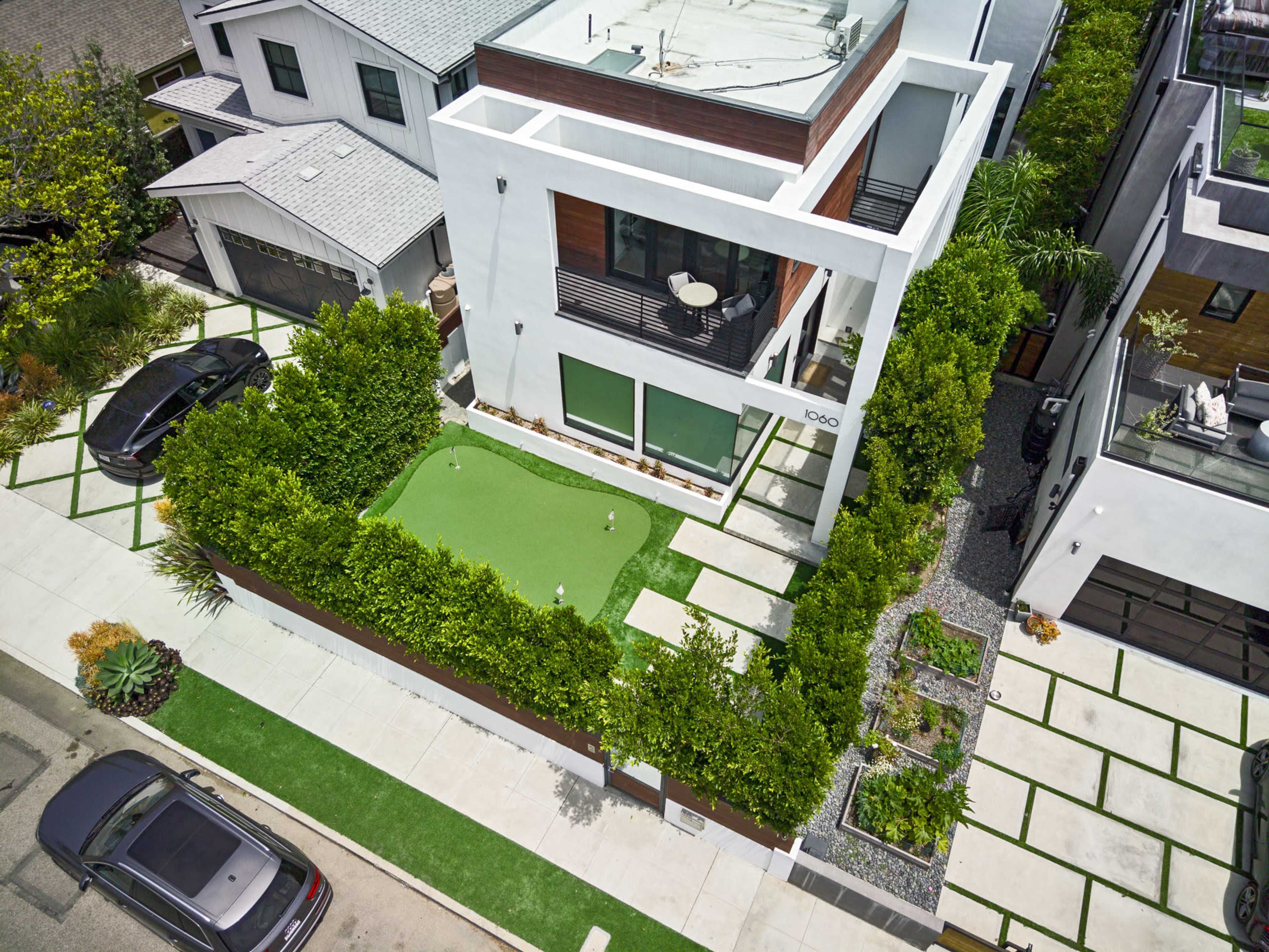 A contemporary house with a landscaped yard featuring artificial turf, surrounded by greenery and walkways, located on a residential street.