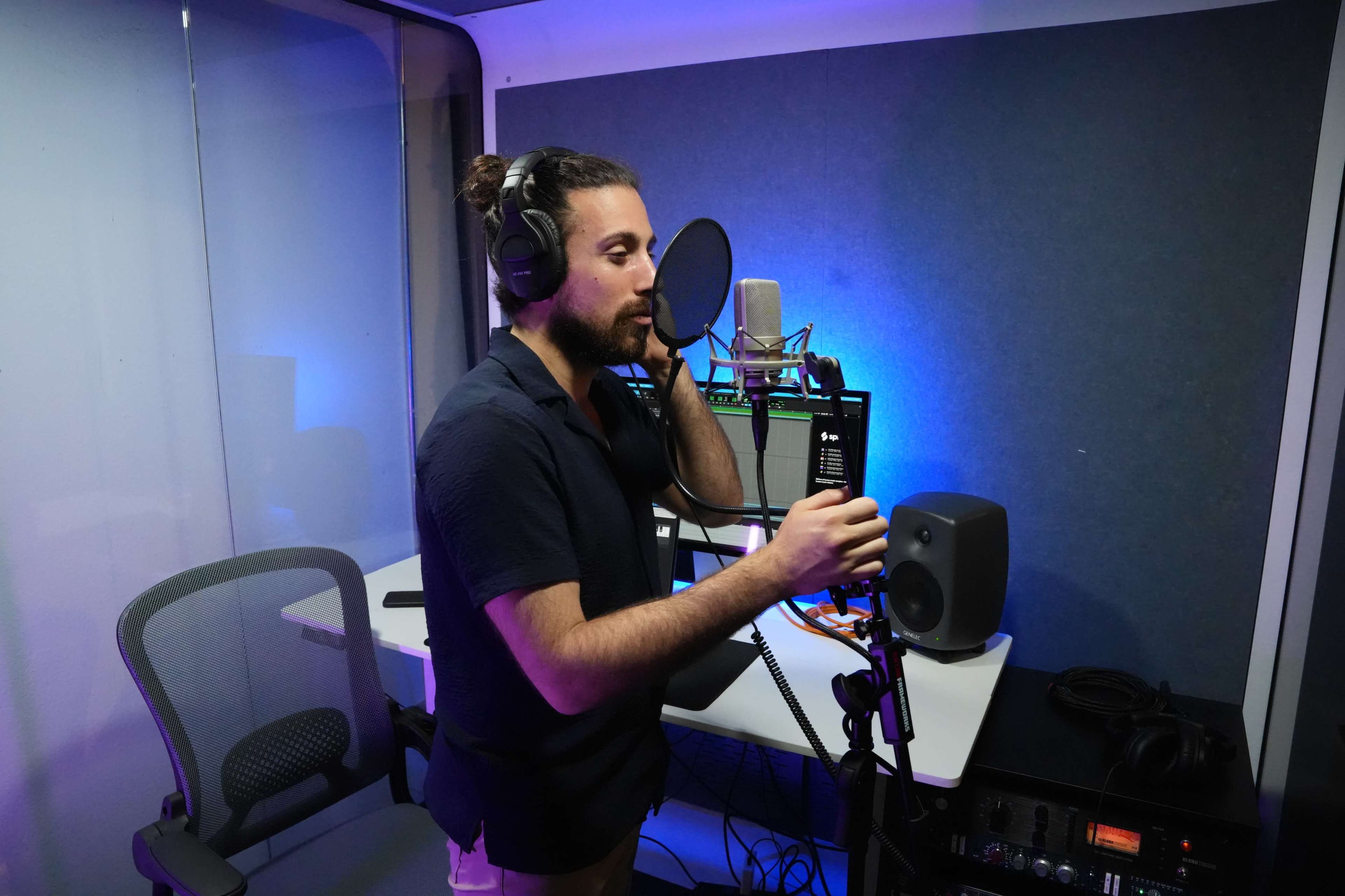 A man is standing in a soundproof recording booth, wearing headphones and speaking into a microphone.