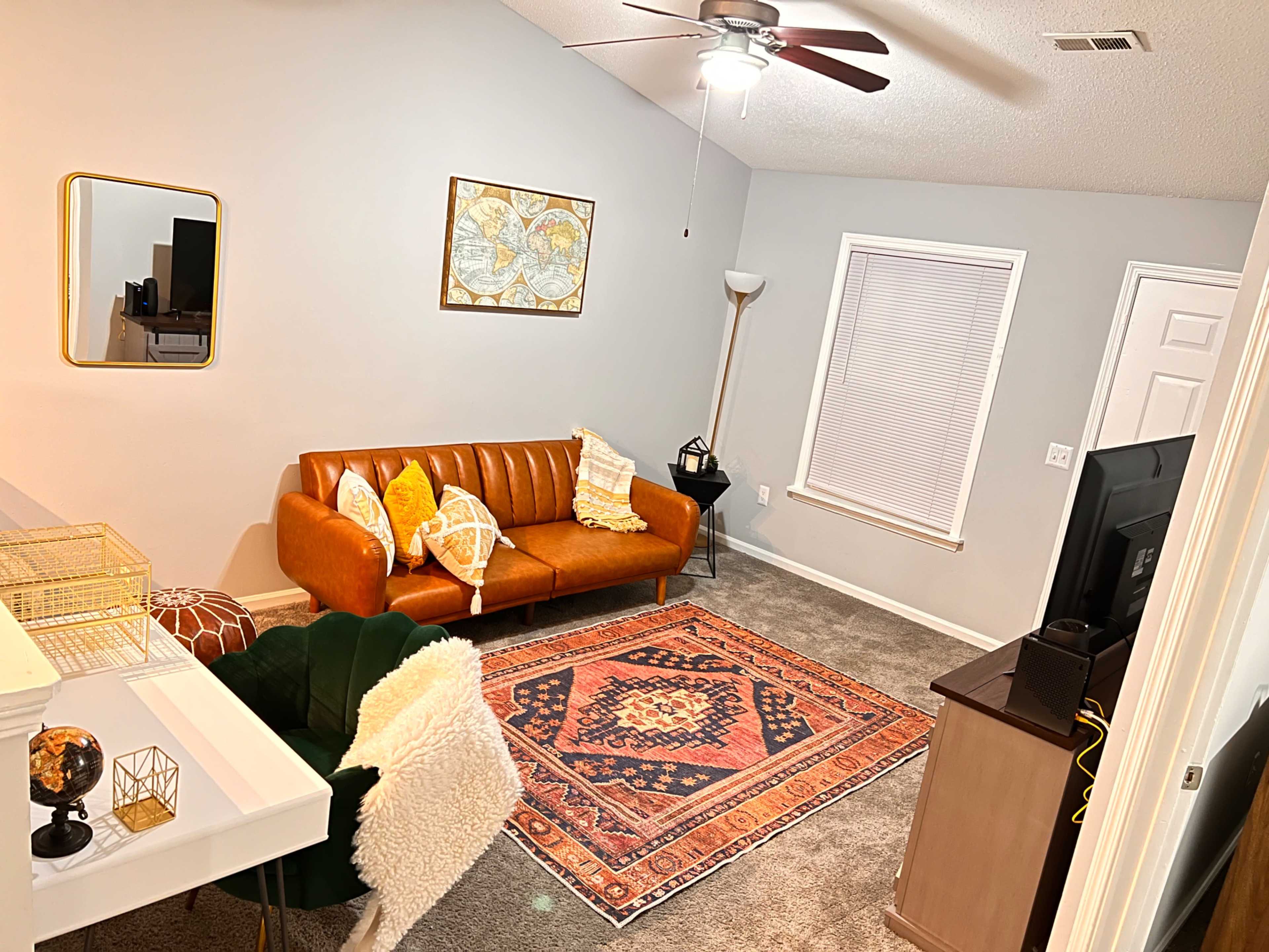 A cozy living room featuring a brown leather sofa, a patterned rug on carpeted flooring, and a desk against the wall.