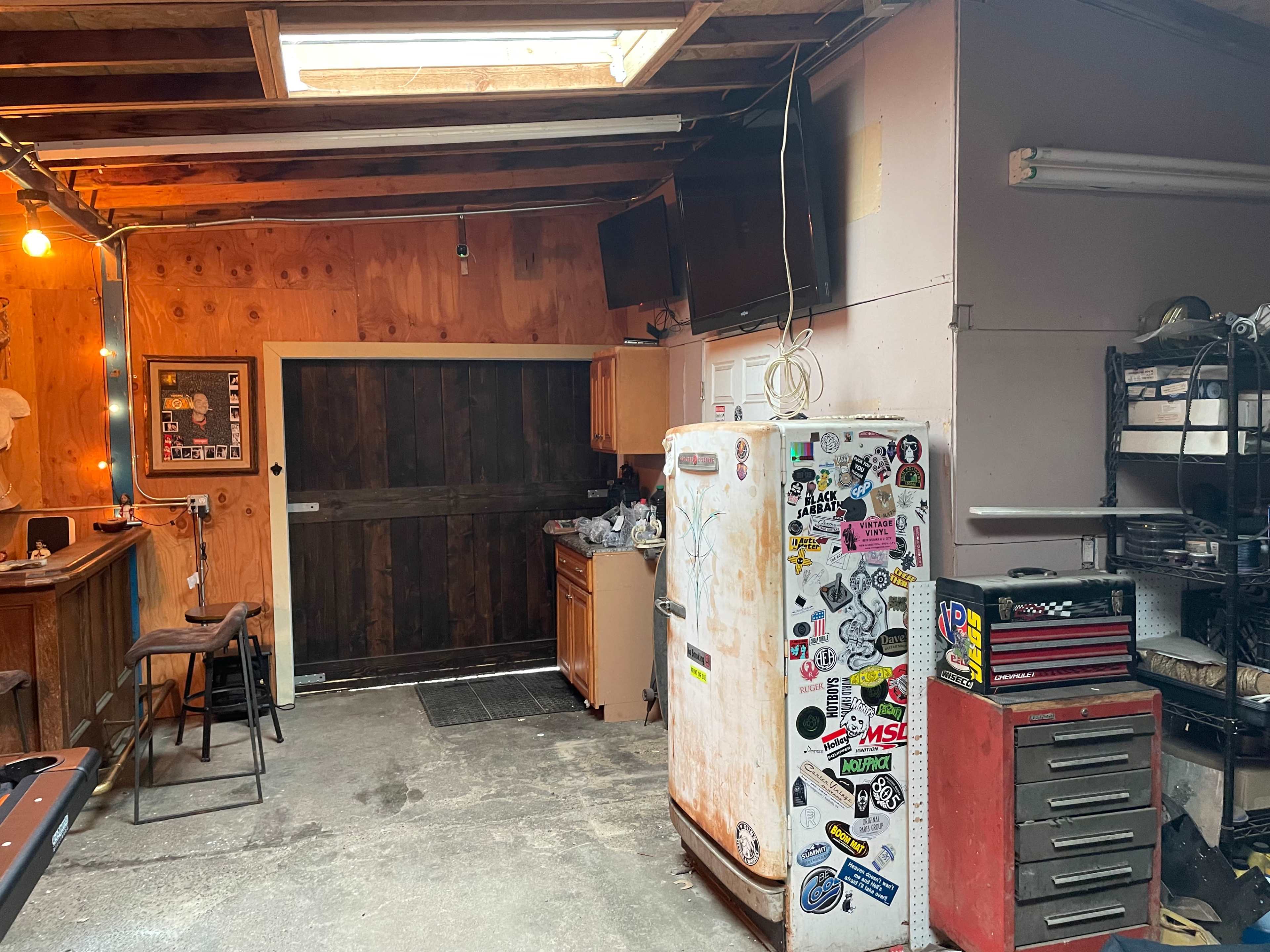 A cluttered garage interior featuring a vintage refrigerator covered in stickers, a tool chest, and a wooden door leading outside.