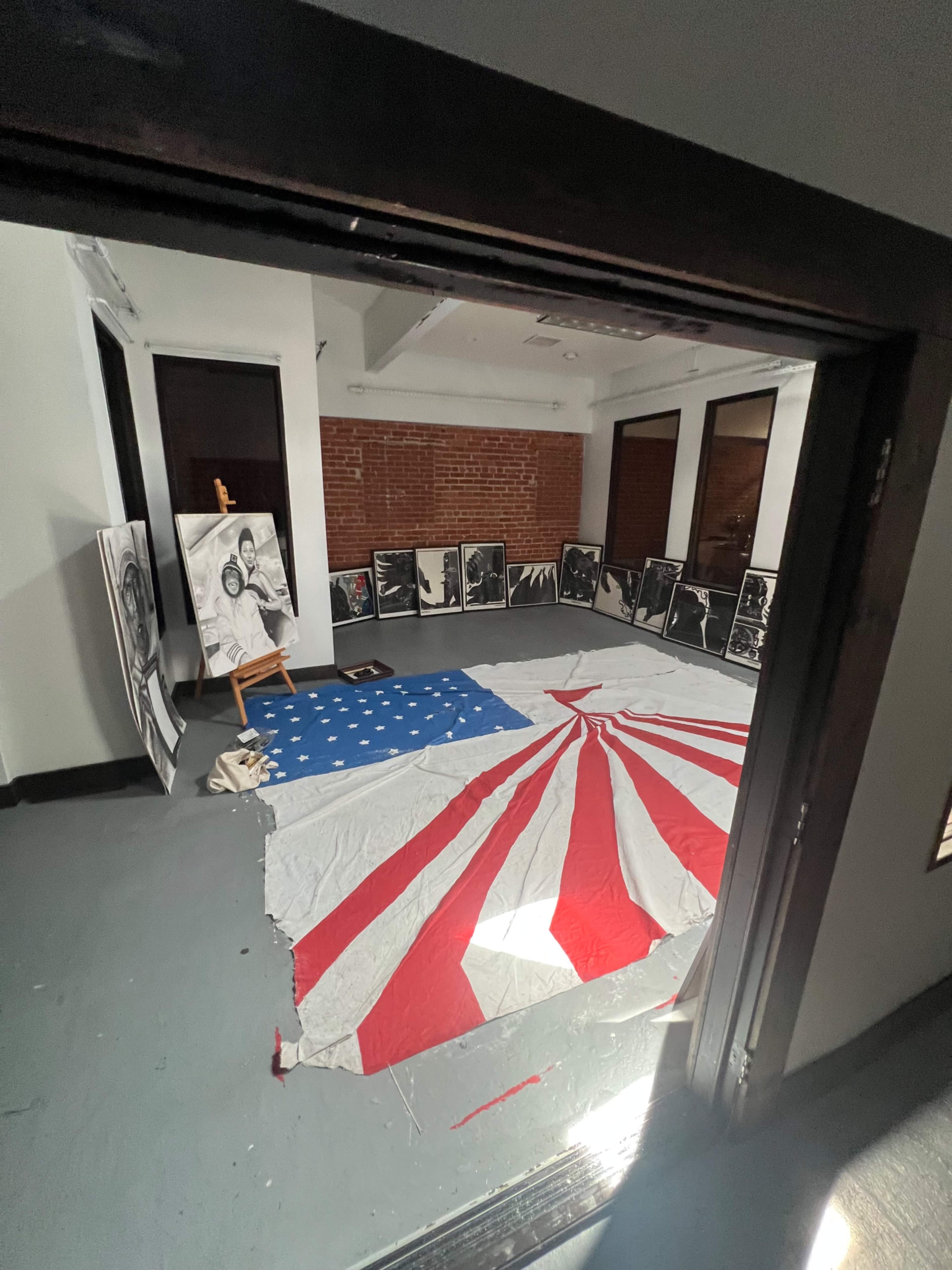 The image shows a room with a large red and white fabric arranged in a starburst pattern on the floor, surrounded by black and white framed photographs leaning against the walls.