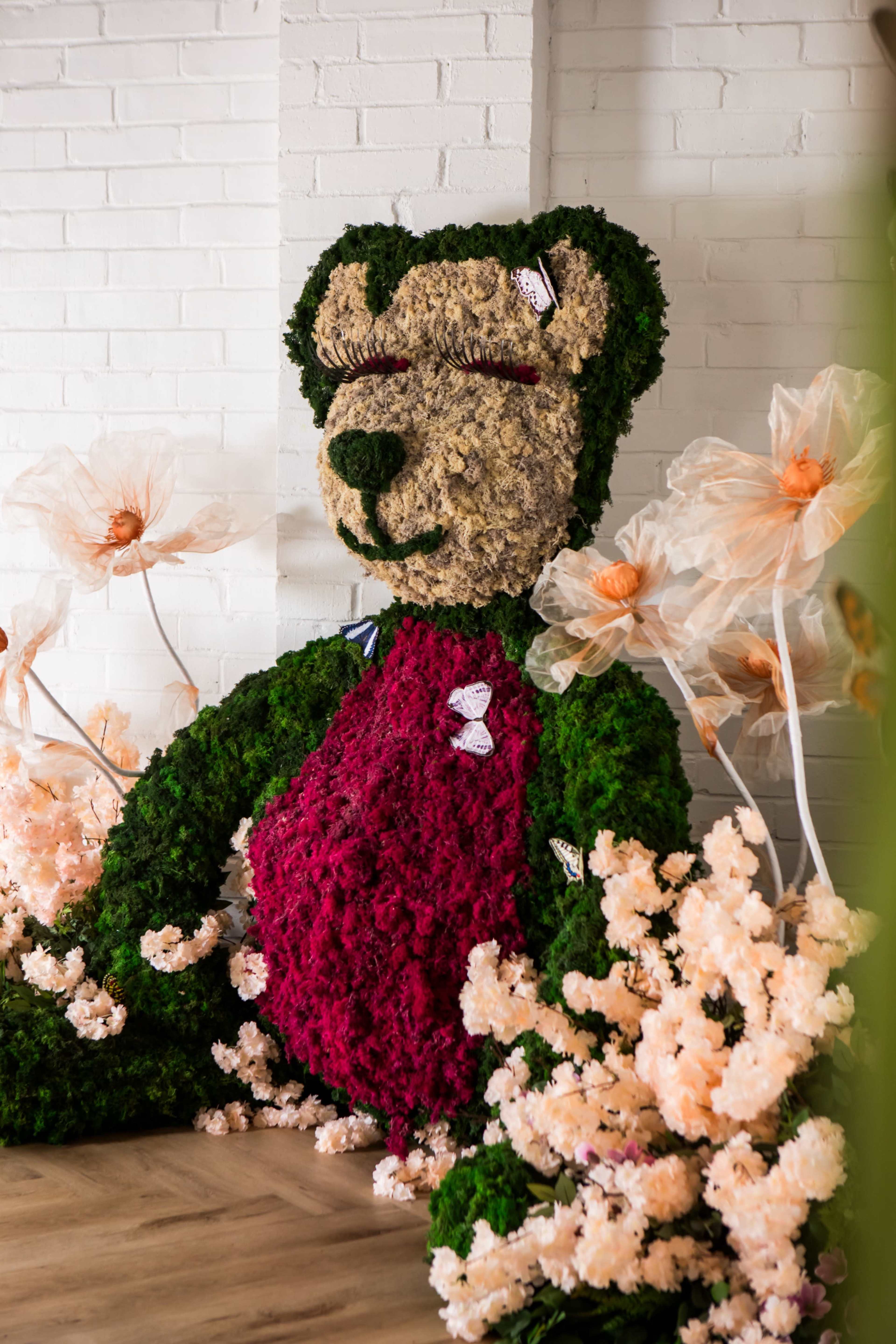A large bear sculpture made of greenery is surrounded by pink and white artificial flowers against a white brick wall.