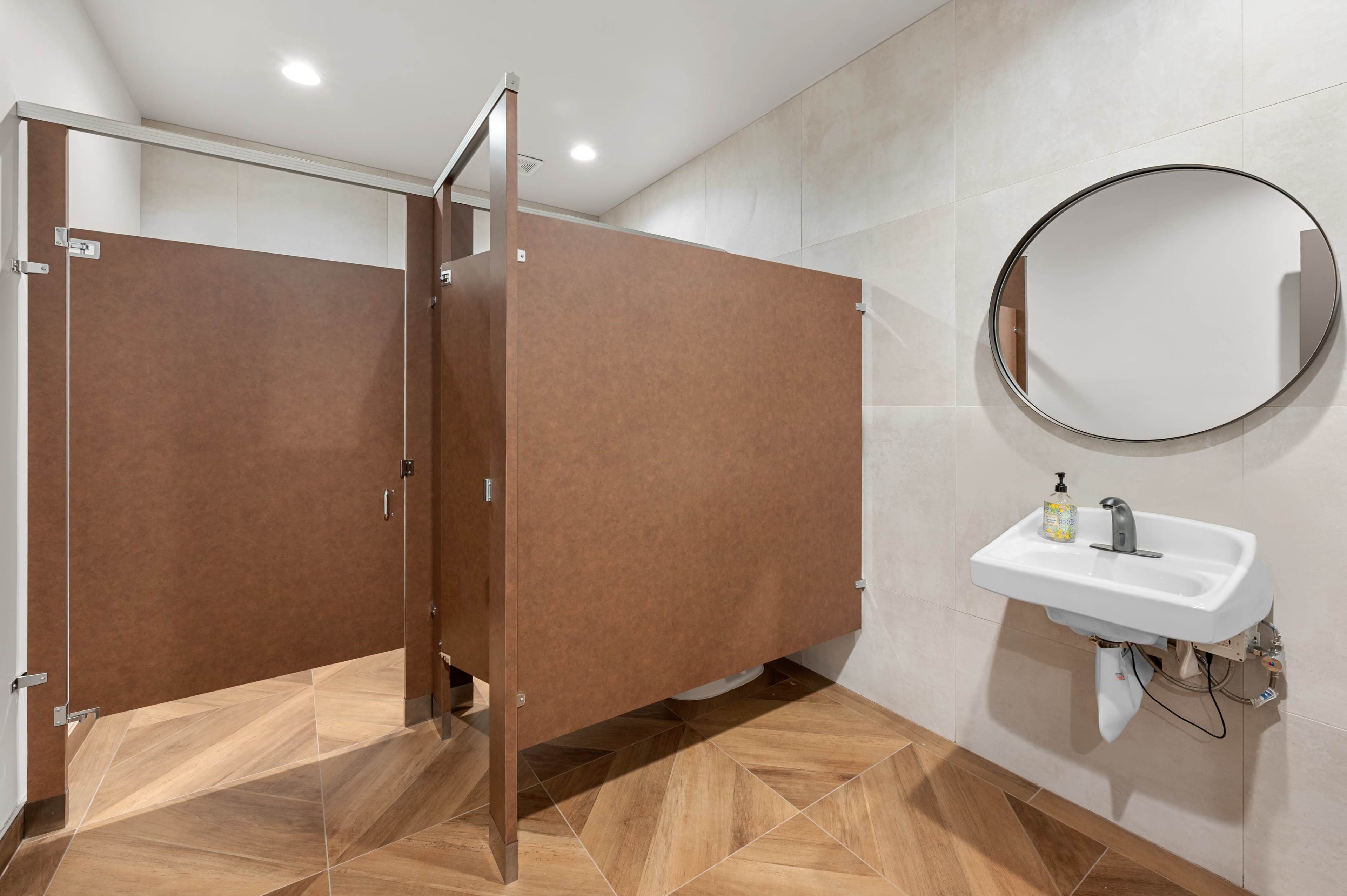 The image shows a modern public restroom with brown partition doors and a circular mirror above a sink.
