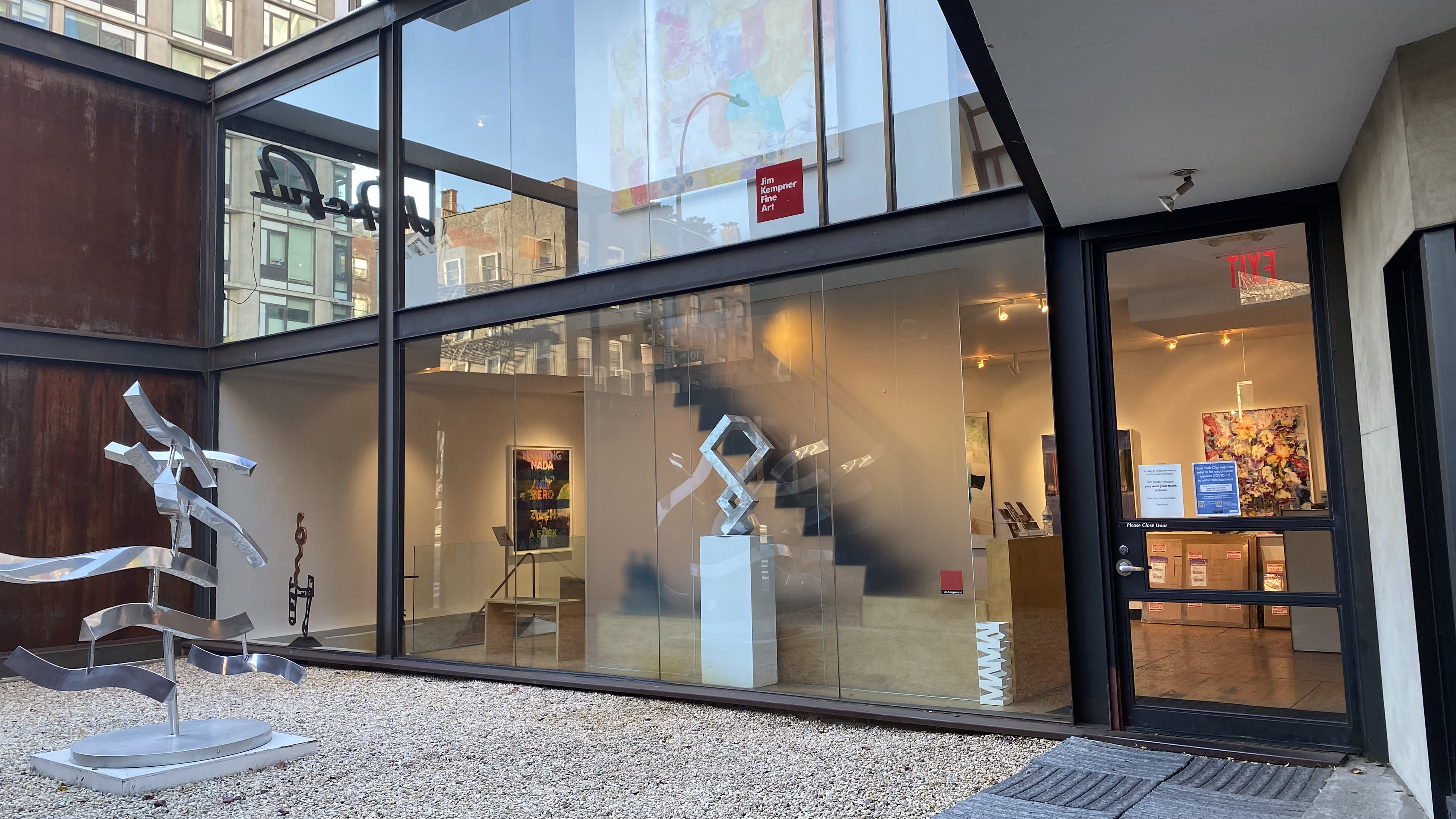 The image shows a modern art gallery featuring a glass facade, a staircase, and a sculpture displayed in the outdoor area.