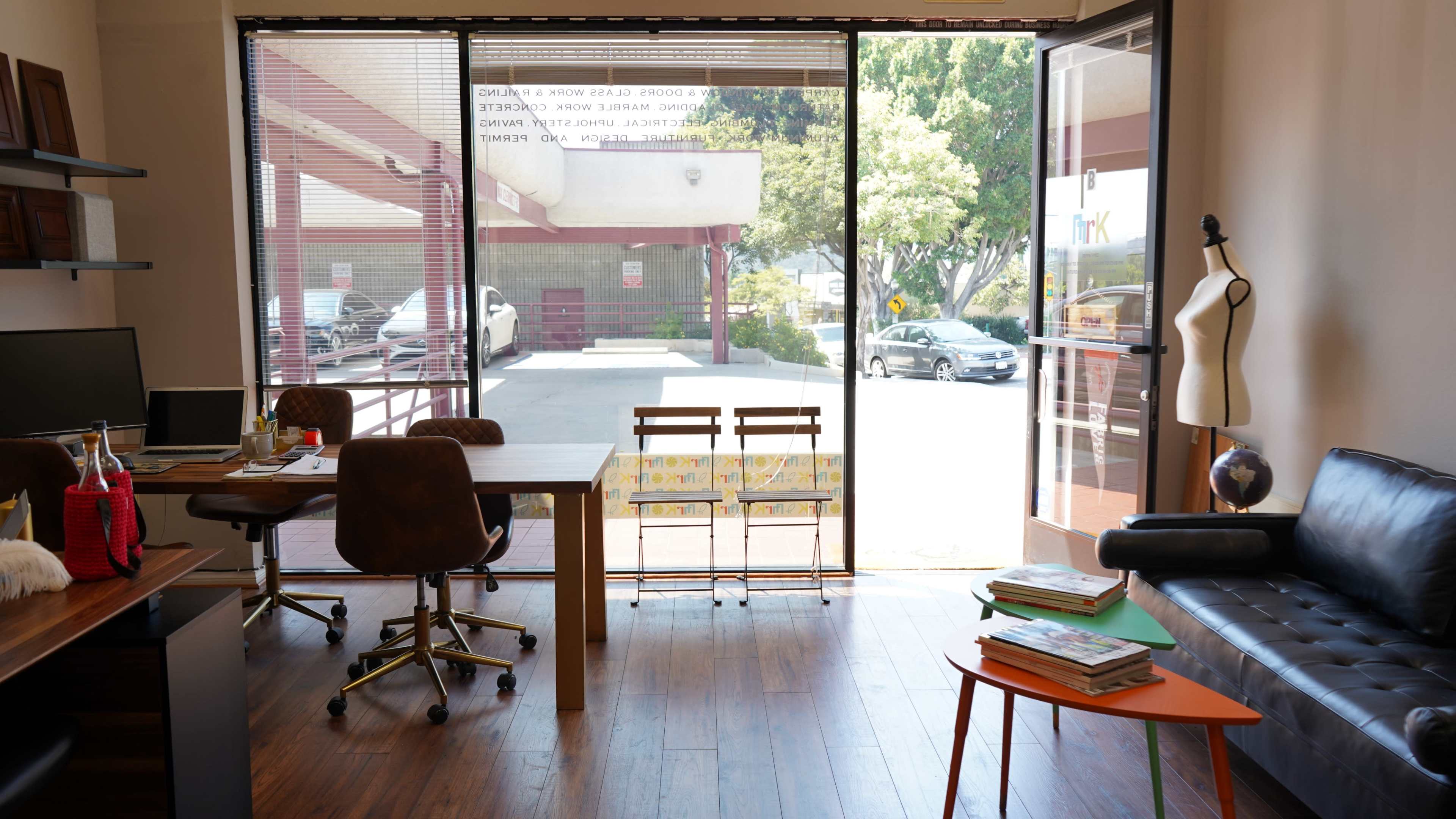 The image shows an office space with a desk, two chairs, a small table, and large windows overlooking a parking area.