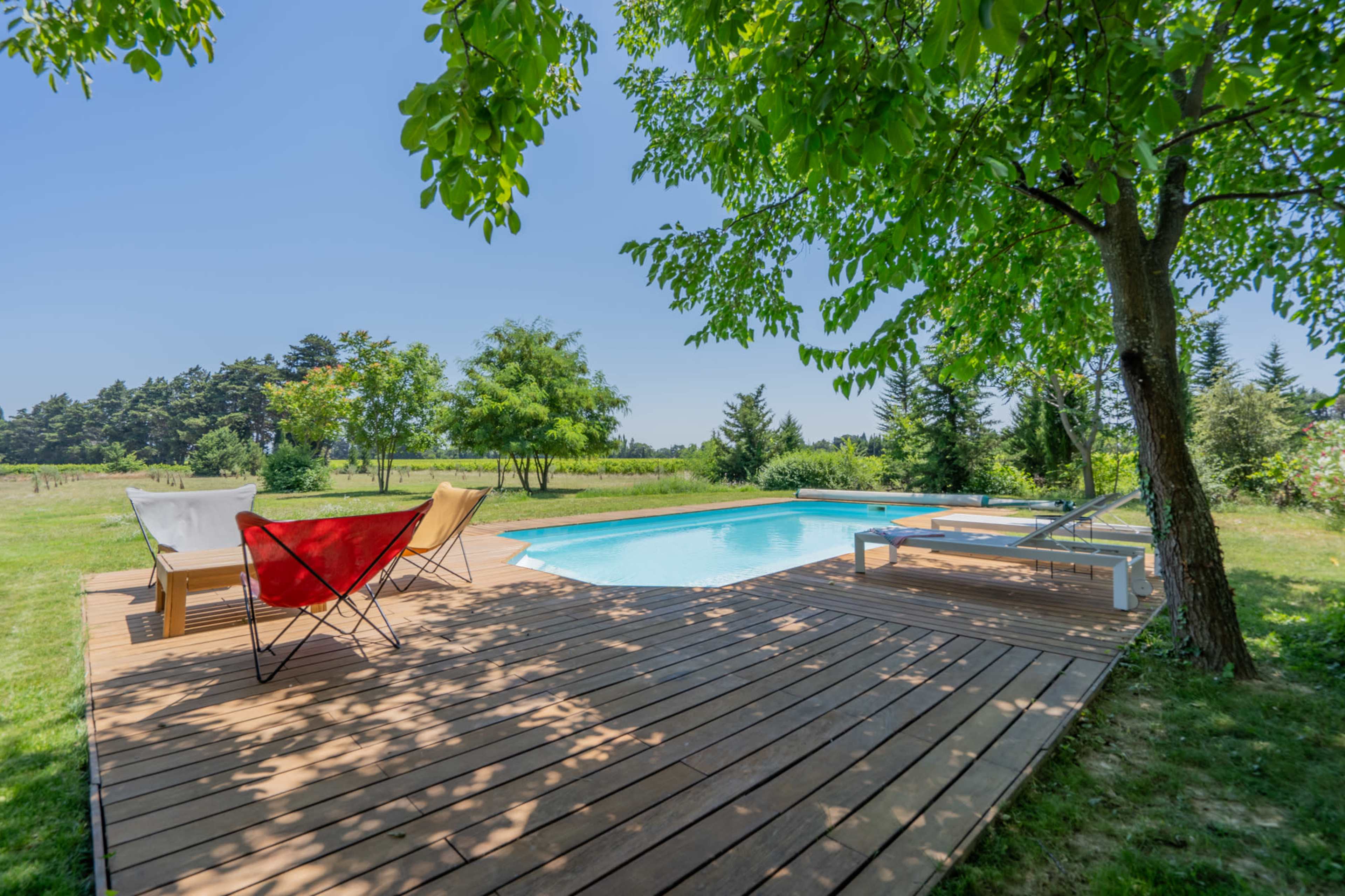 A wooden deck with lounge chairs surrounds a swimming pool amidst a grassy landscape with trees in the background.