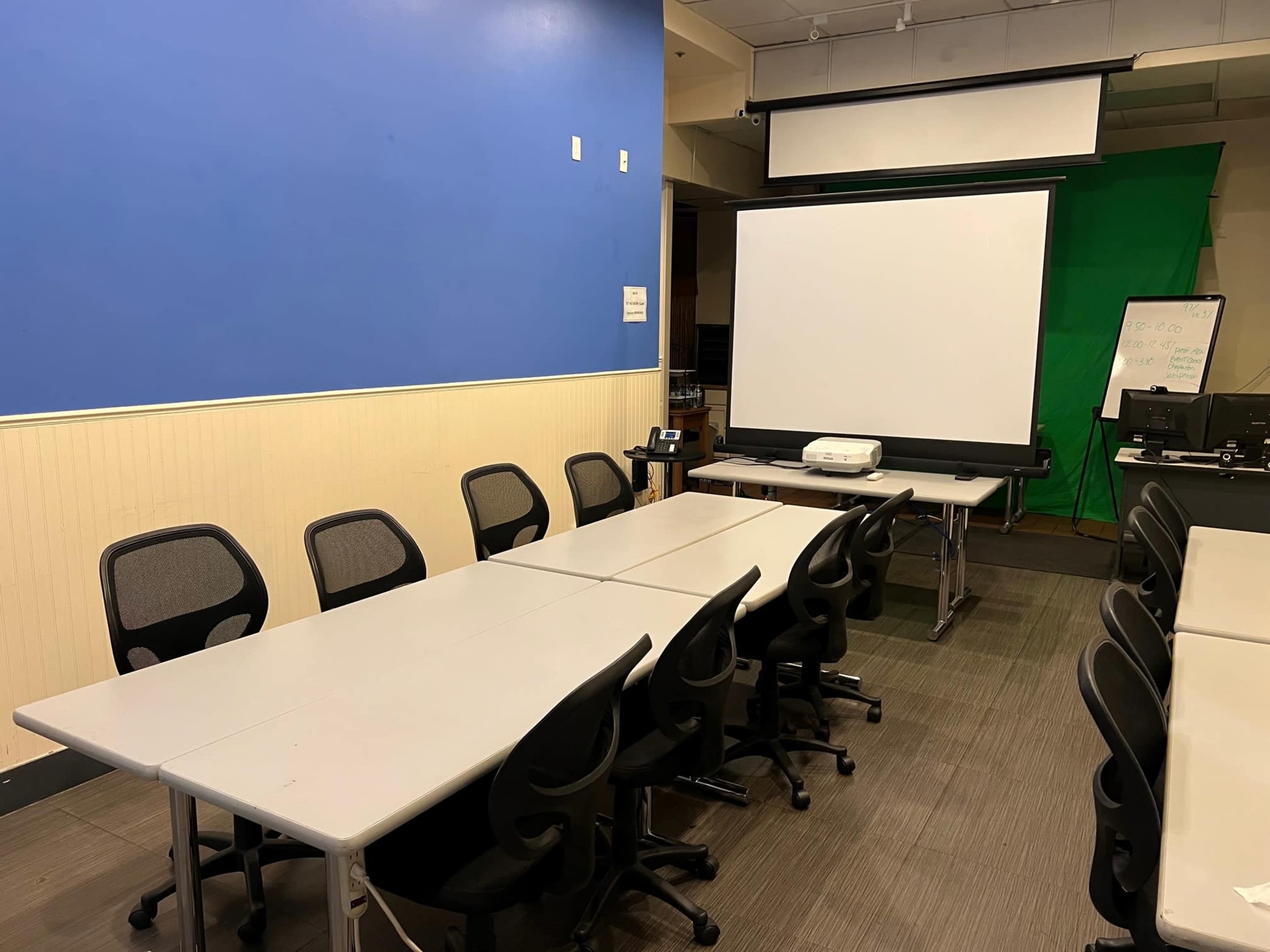 A classroom features a long table with several black chairs, a projector on a table, and a large screen against a blue wall.