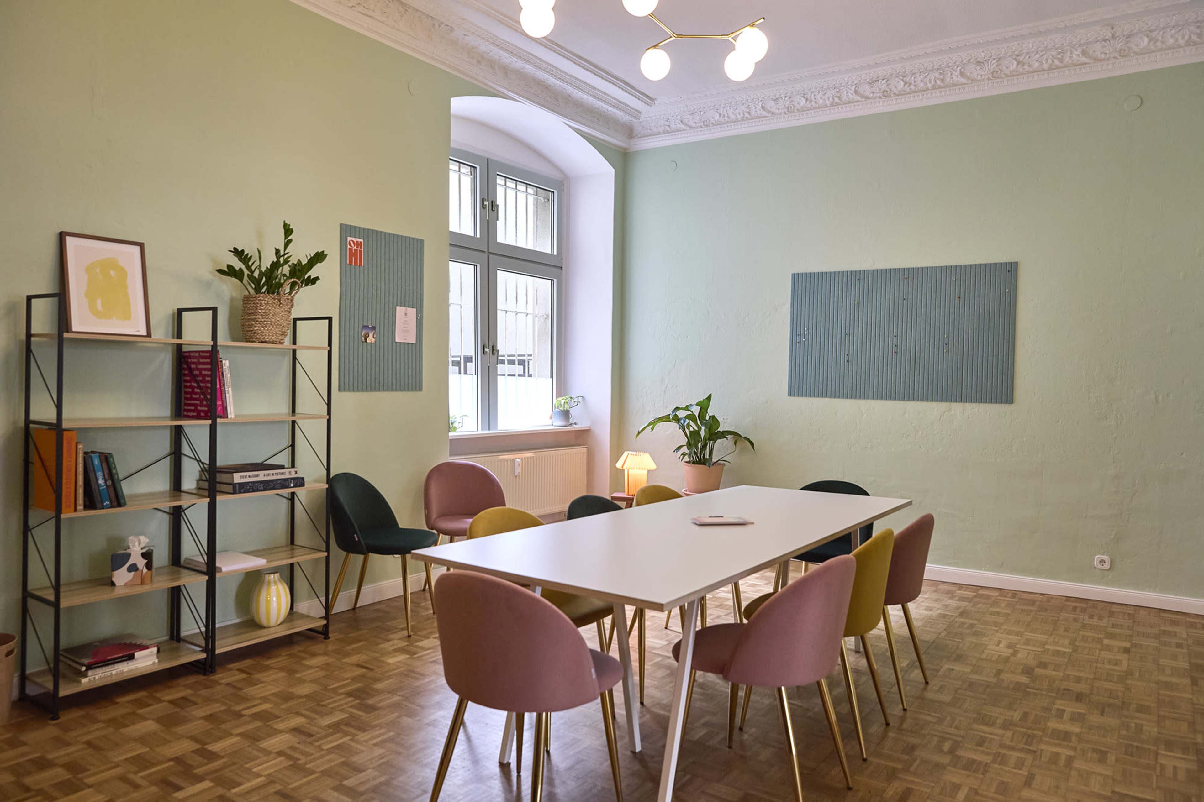 The image shows a light green room with a long white table surrounded by multi-colored chairs, a bookshelf with decorative items, and a window letting in natural light.