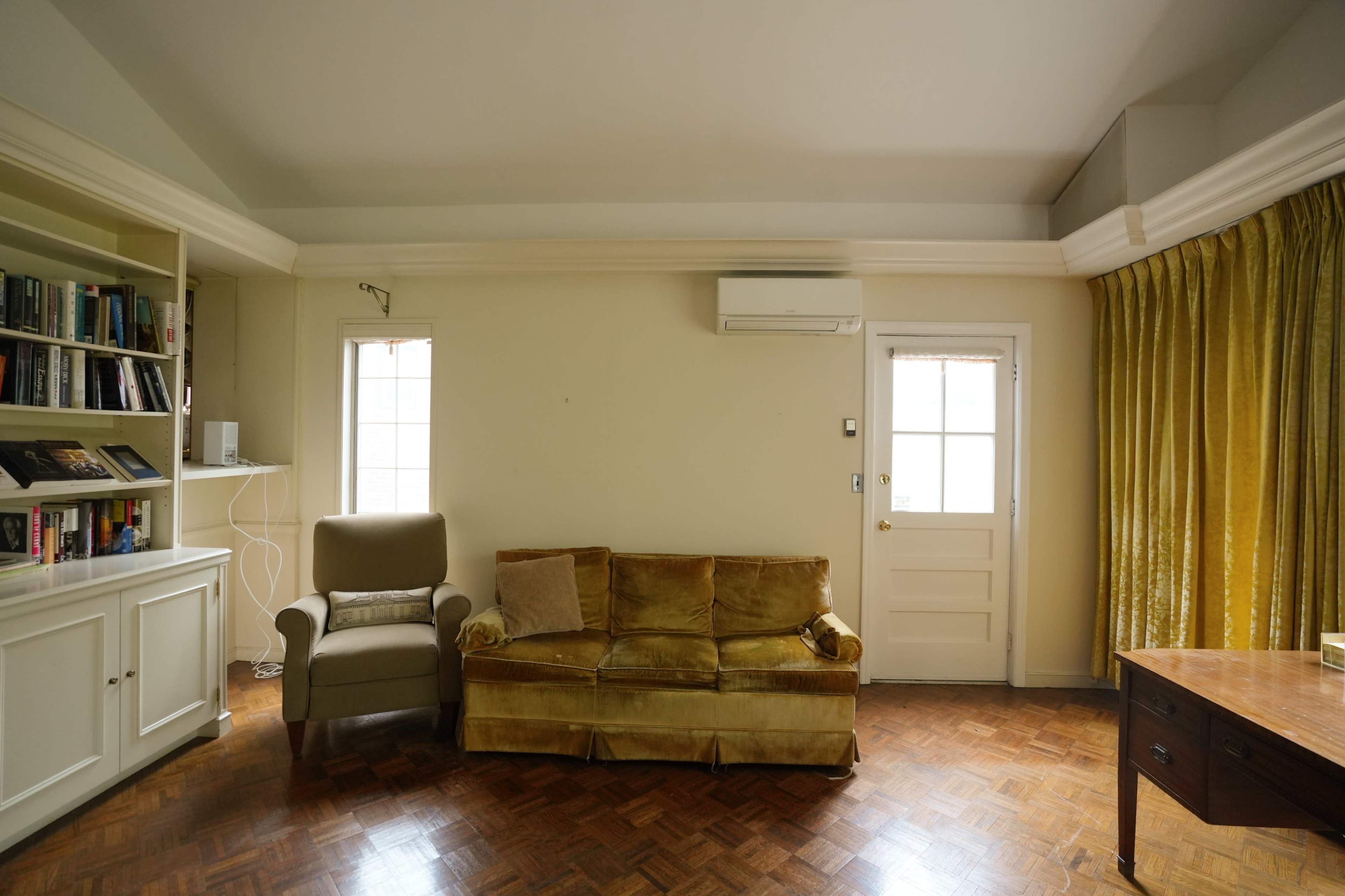 The room features a gold velvet sofa, an armchair, a wooden table, a bookshelf, and large windows with yellow curtains.