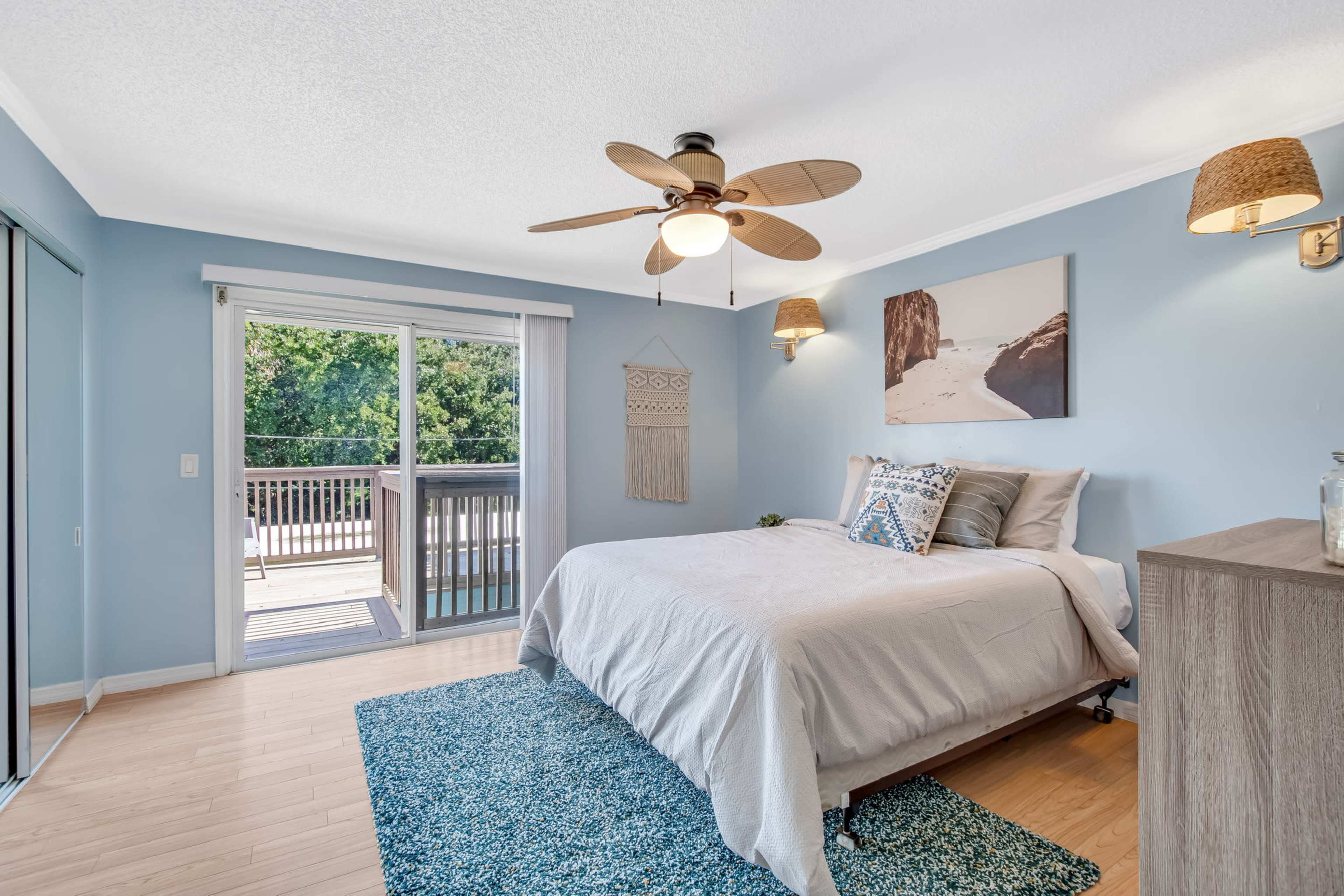 A bedroom with a bed, a ceiling fan, and sliding glass doors leading to a deck surrounded by trees.